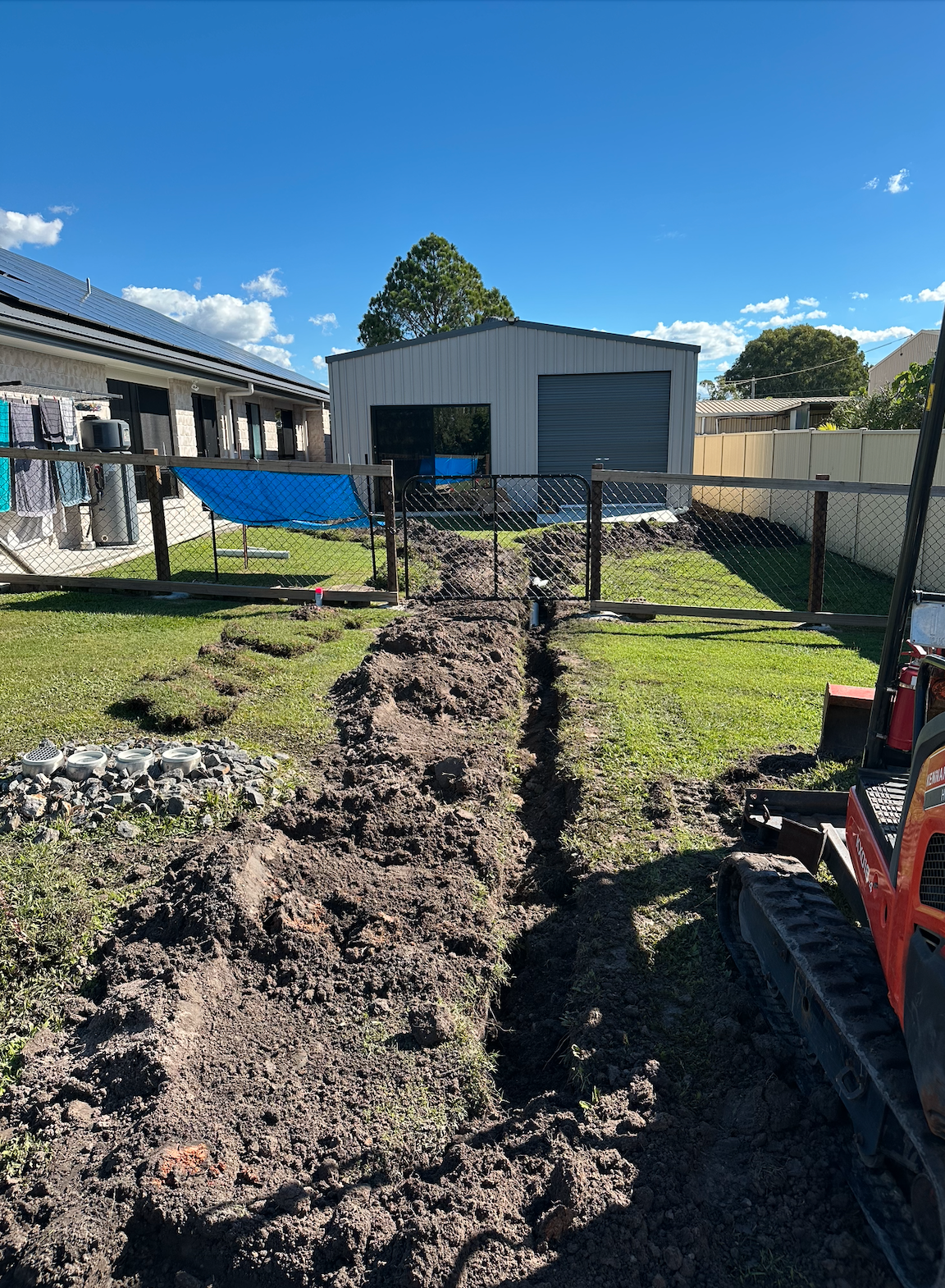 A backyard construction site with a trench dug along the grass, a small excavator parked on the right, and a fenced yard with houses and a shed in the background under a blue sky with clouds.