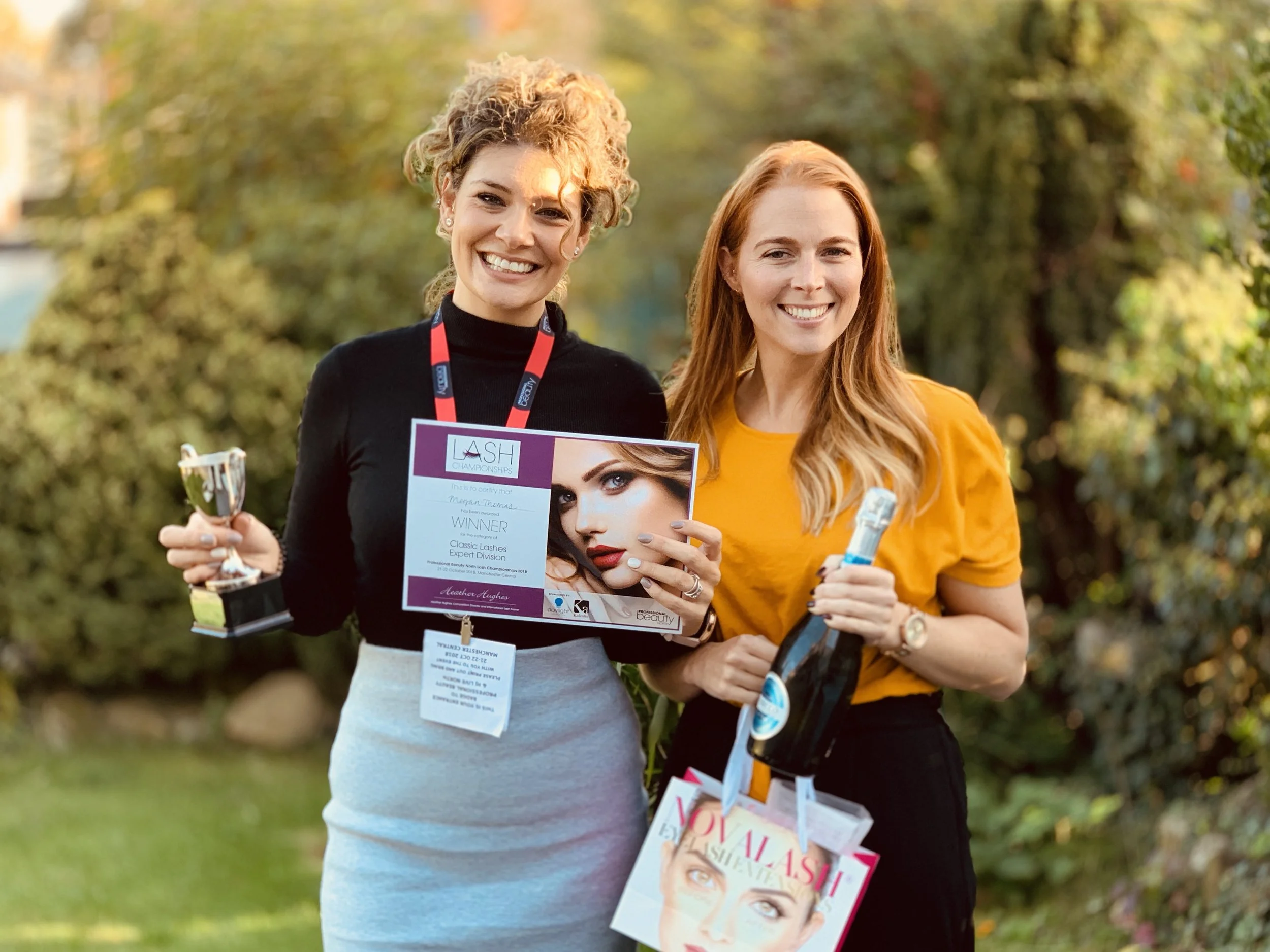 Two women smiling outdoors holding awards and magazines, with trees in the background. Megan Thomas Lash Academy