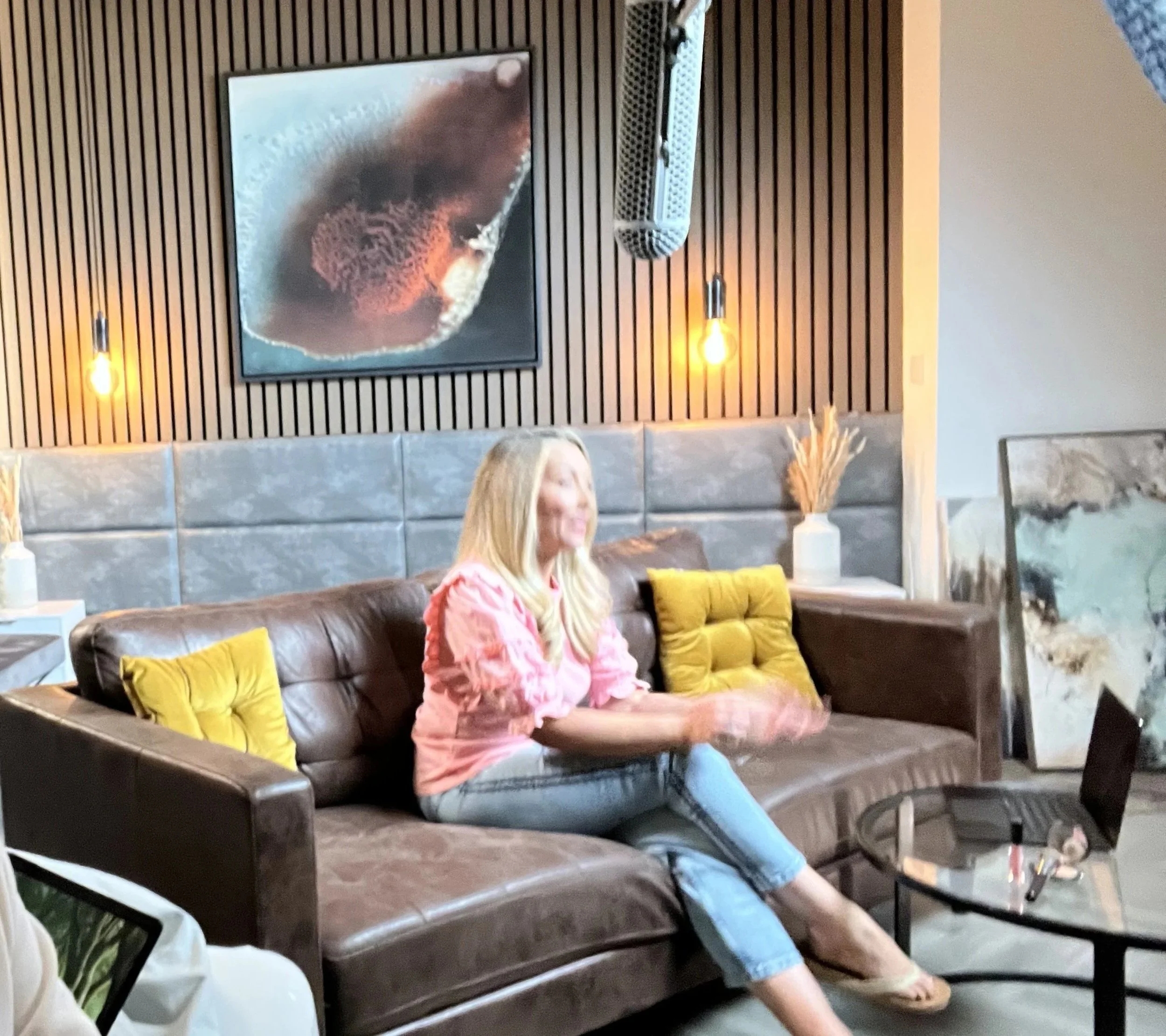 A woman in a pink top and jeans sitting on a brown sofa with yellow pillows in a modern living room with wooden wall panels and wall-mounted lights, a large abstract painting, a side table with decorative vases, and a glass coffee table with a laptop and objects.