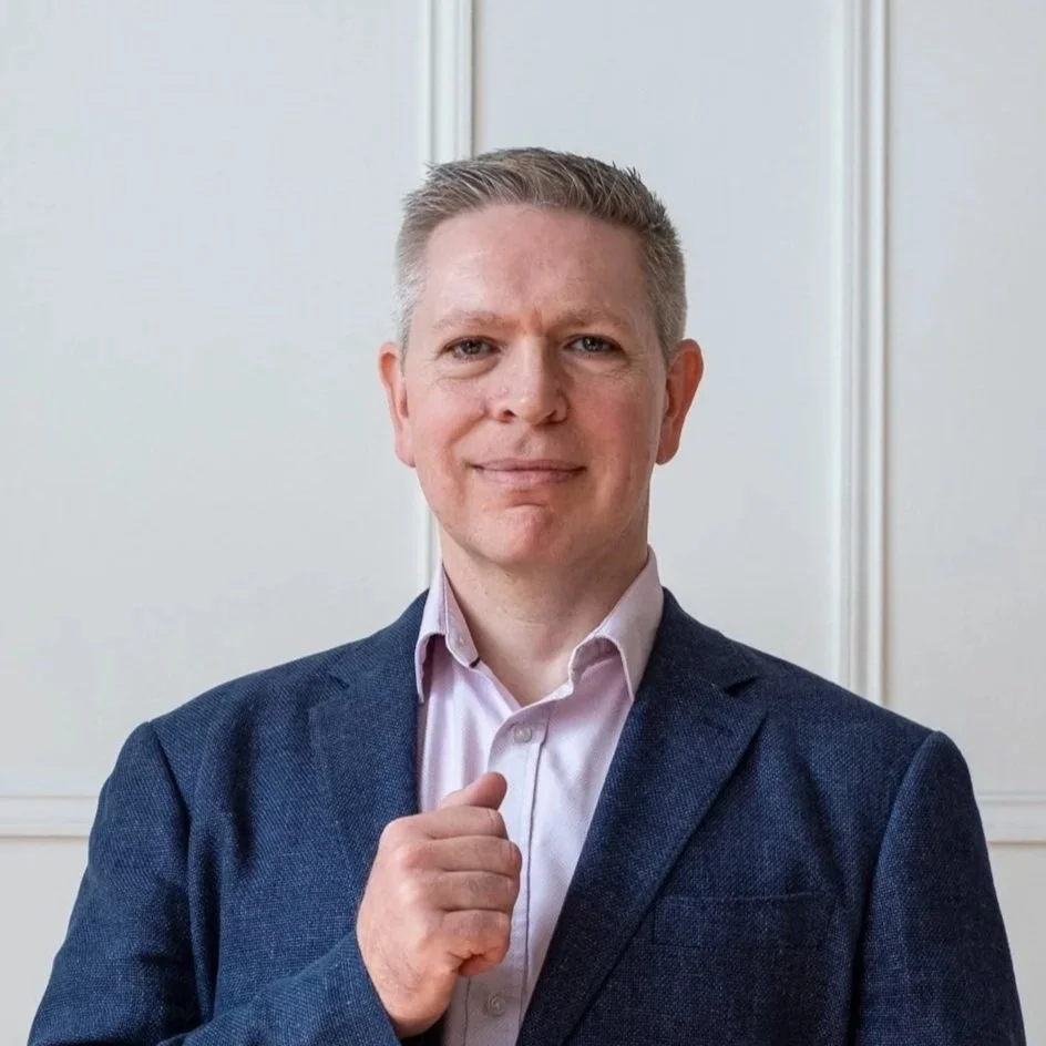 A man in a suit pointing to himself with a confident expression.