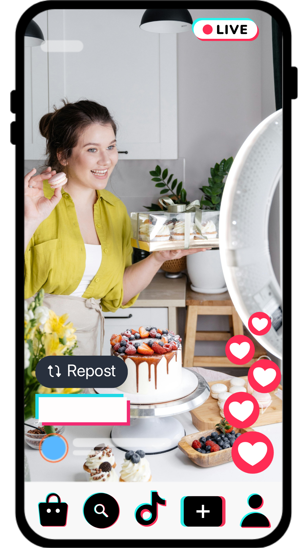 A woman in a yellow shirt and apron is filming a baking video in a kitchen, holding a box of desserts, with a decorated cake and various pastries and berries on the table in front of her from Live Sell Academy