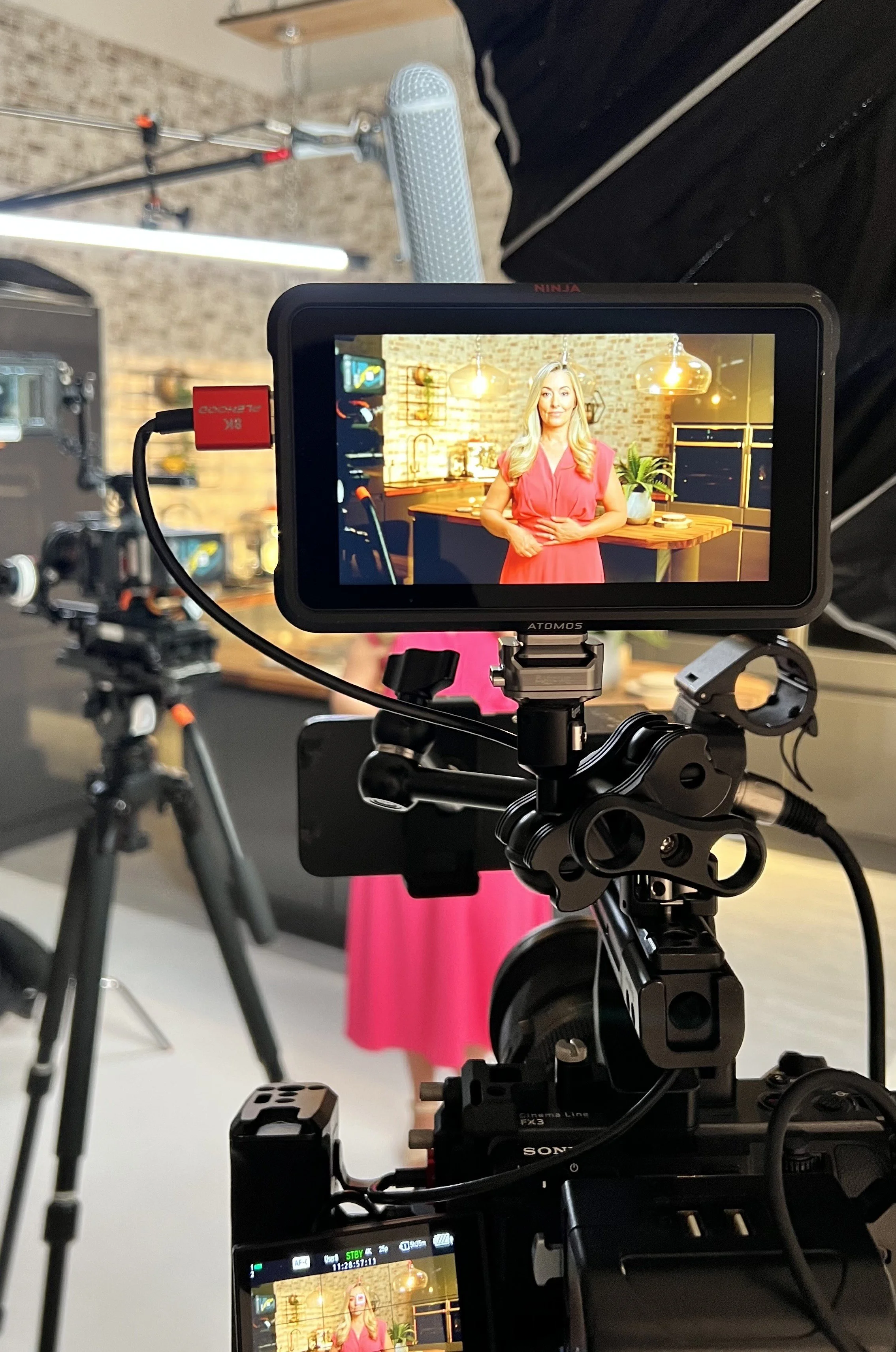 A video camera recording a woman with blonde hair in a pink dress standing in a modern kitchen set with brick walls and warm lighting.