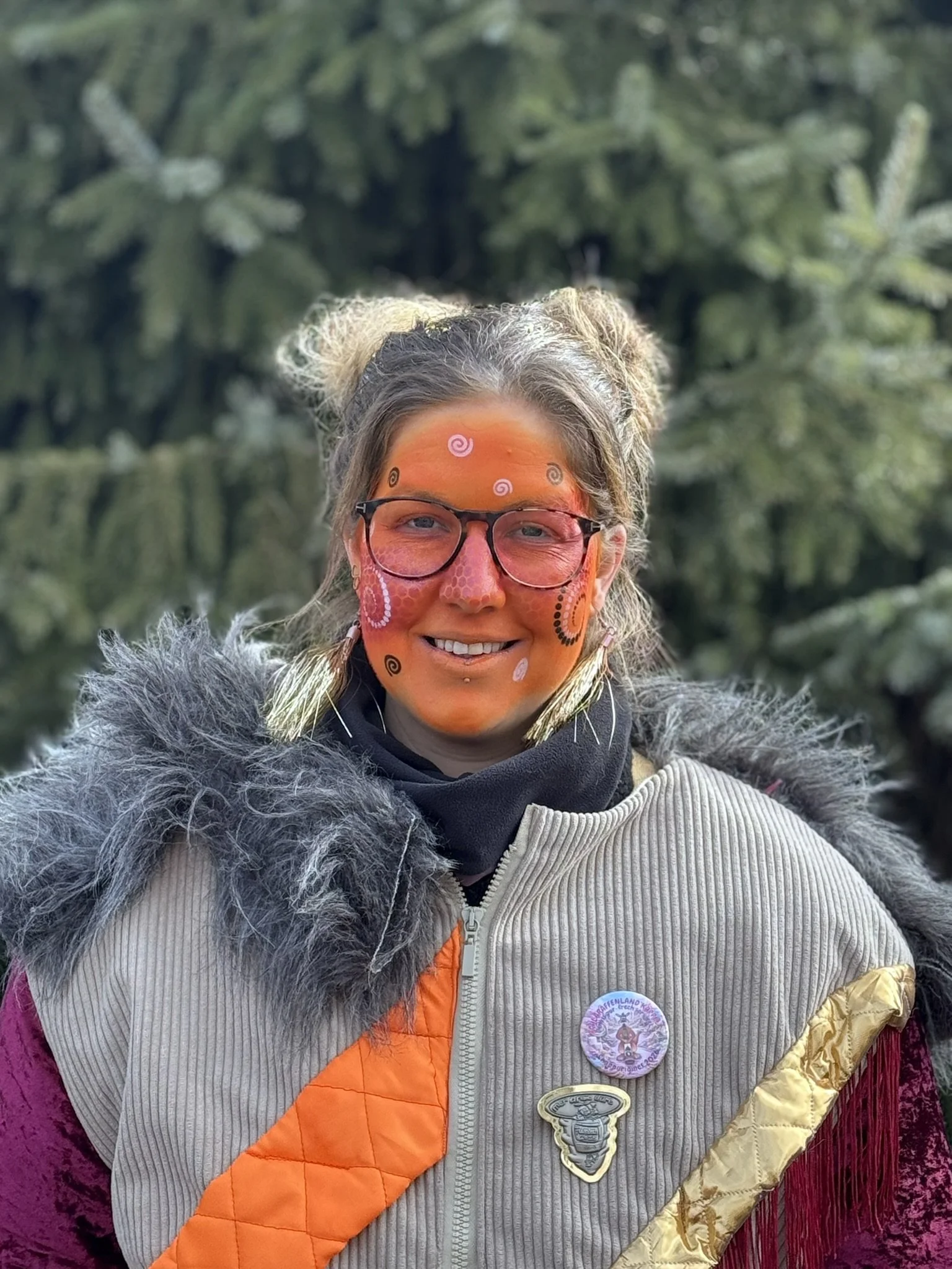 Junge Frau mit bunten Gesichtsmalereien, trägt Brille, Mütze mit Text, schwarze Jacke mit Pins, im Freien vor grünen Büschen.