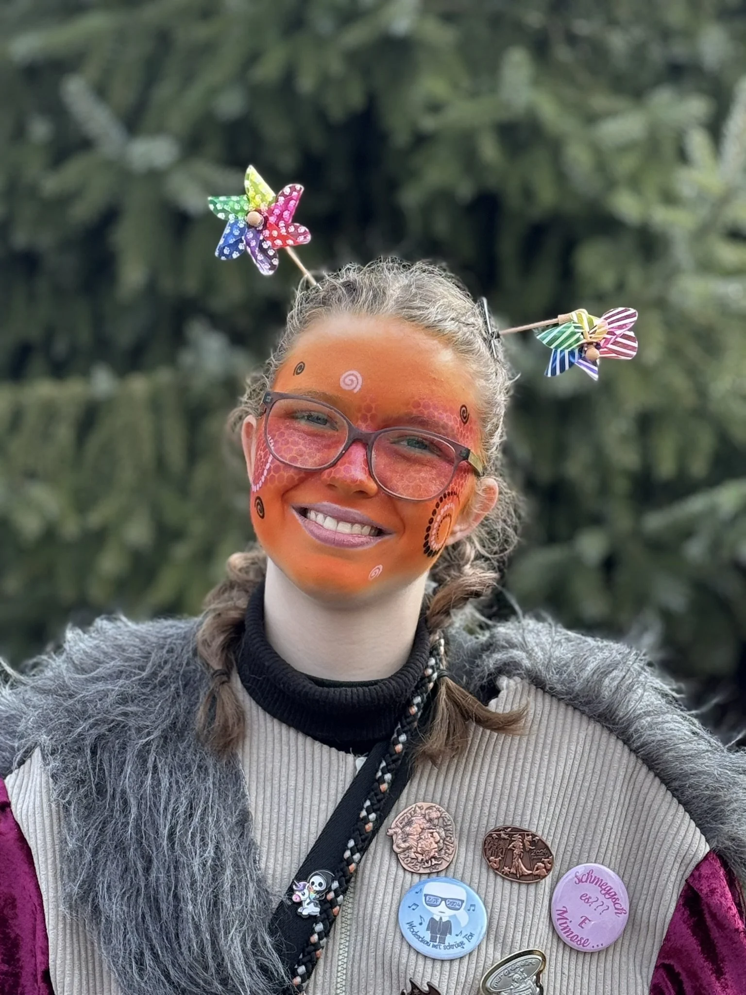 Junge Frau mit Gesichtsbemalung in Rot und Blau, trägt Brille, schwarze Jacke mit Pins, steht vor grünen Büschen und lächelt.