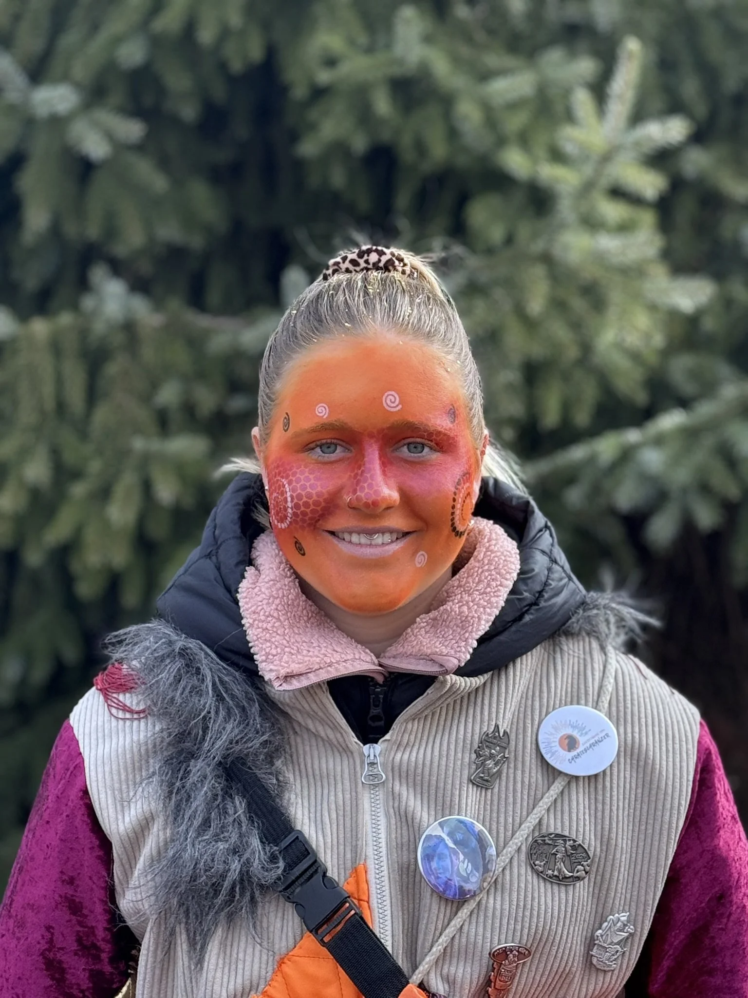 Junge Frau mit Gesichtsbemalung, Pins und dunklem Jacke, stehend vor grüner Natur im Freien.