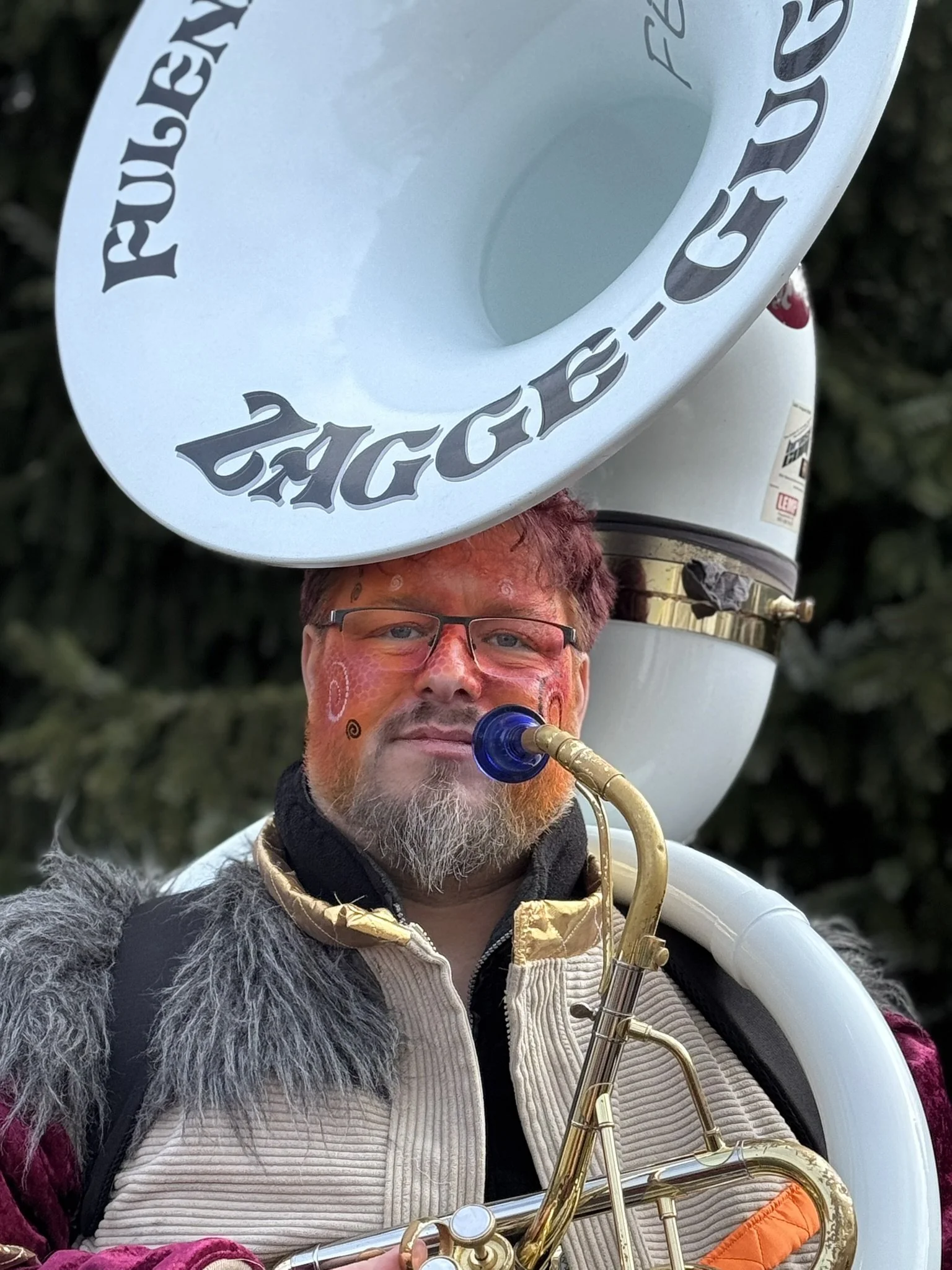 Porträt eines Mannes mit Bart, Brille, grauem Mützen und dunklem Jacke, im Freien vor grünen Büschen an sonnigem Tag.