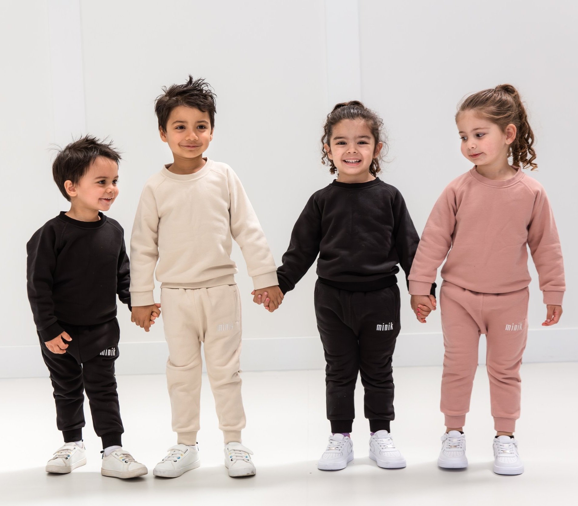 Four children standing in a row, wearing matching outfits in black, beige, and pink, holding hands and smiling. They are indoors on a white background.