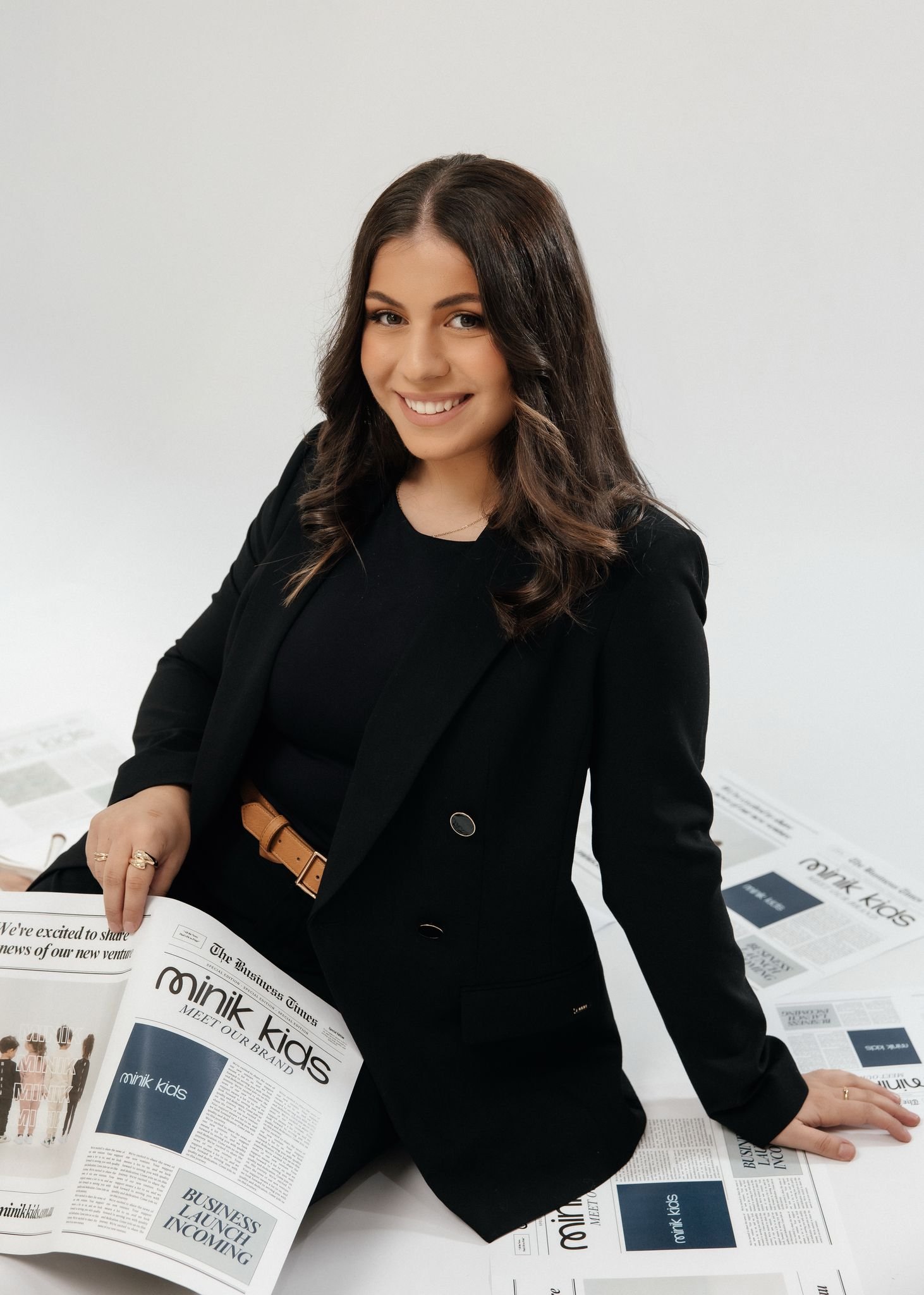 The designer Ayshe is smiling while sitting on a floor covered with newspapers titled "minik kids" and "The Business Times."