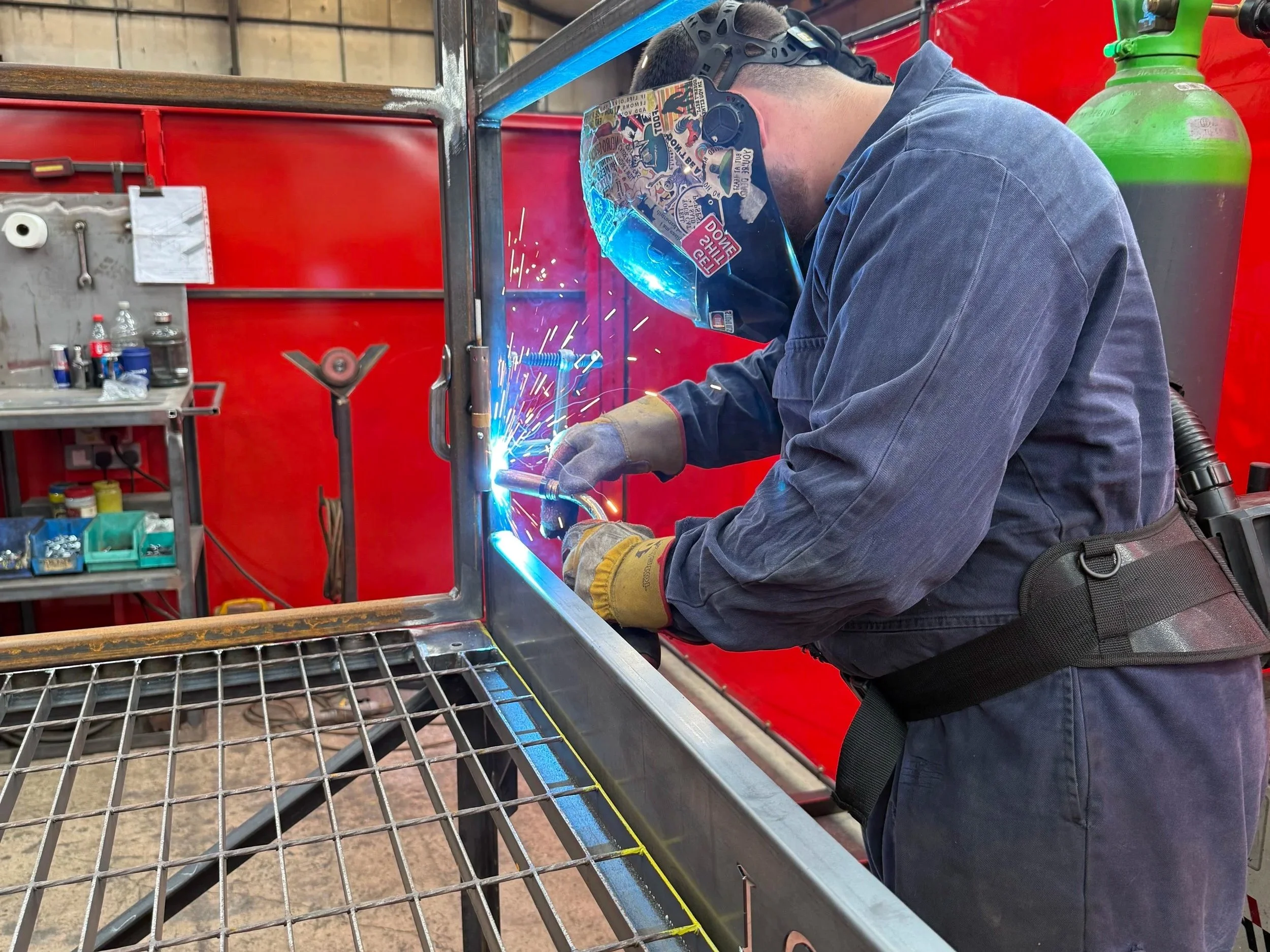 A person wearing a colorful welding helmet and protective gloves is welding two metal pieces together in a workshop. Sparks fly from the welding process, and there are shelves with tools and supplies in the background.