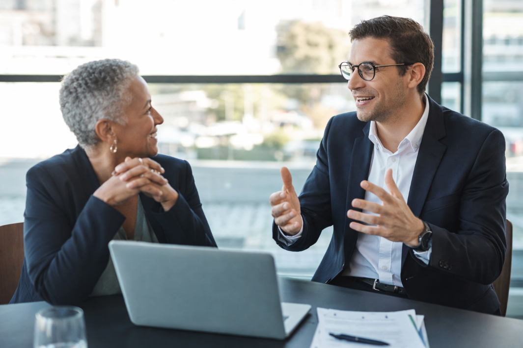 Deux personnes d'affaires discutant lors d'une réunion, une femme plus âgée et un homme plus jeune, dans une salle lumineuse avec une vue urbaine en arrière-plan.