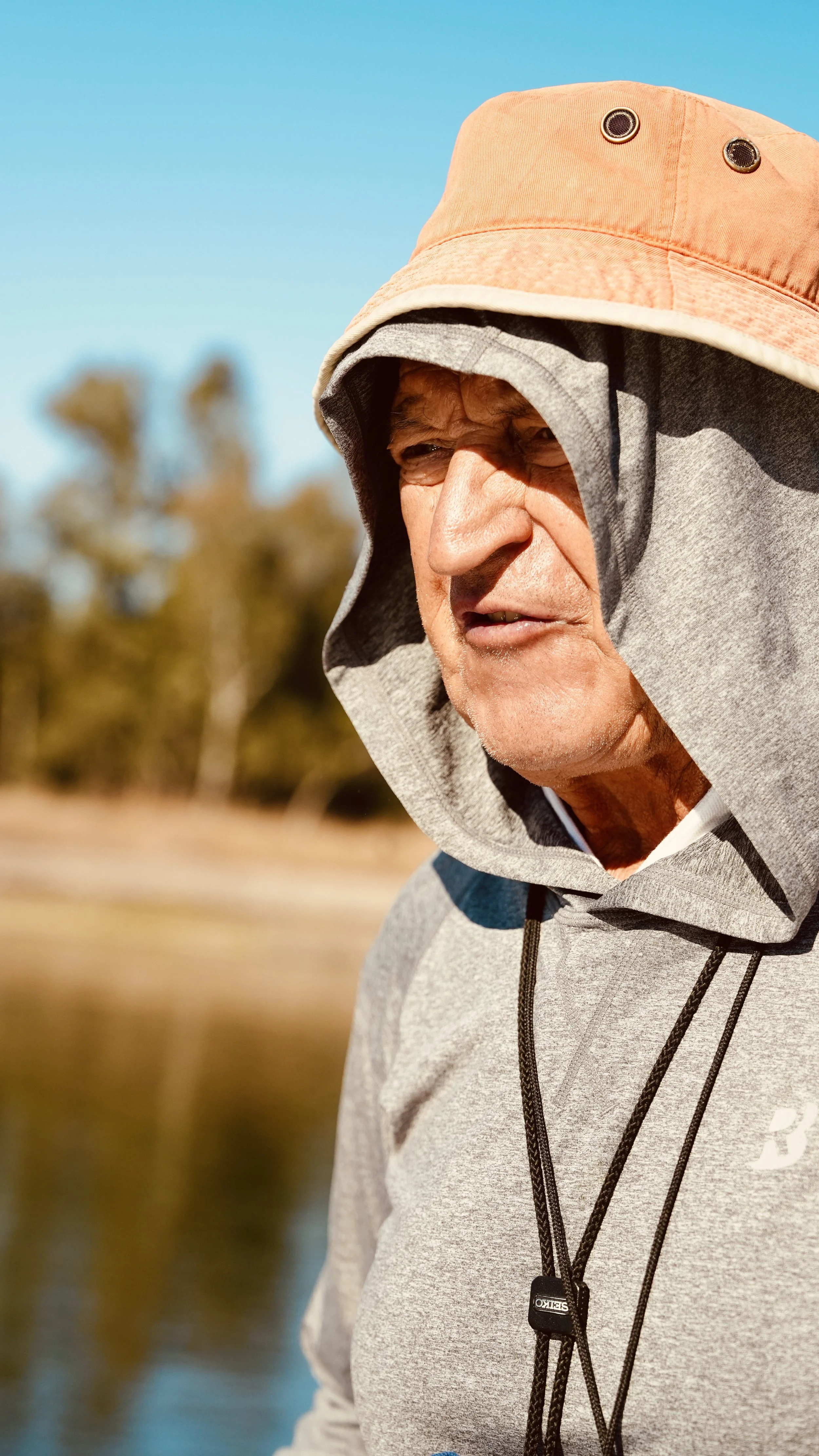 Close-up of an elderly man outdoors wearing a beige wide-brim hat, a hooded gray jacket, and a whistle around his neck.