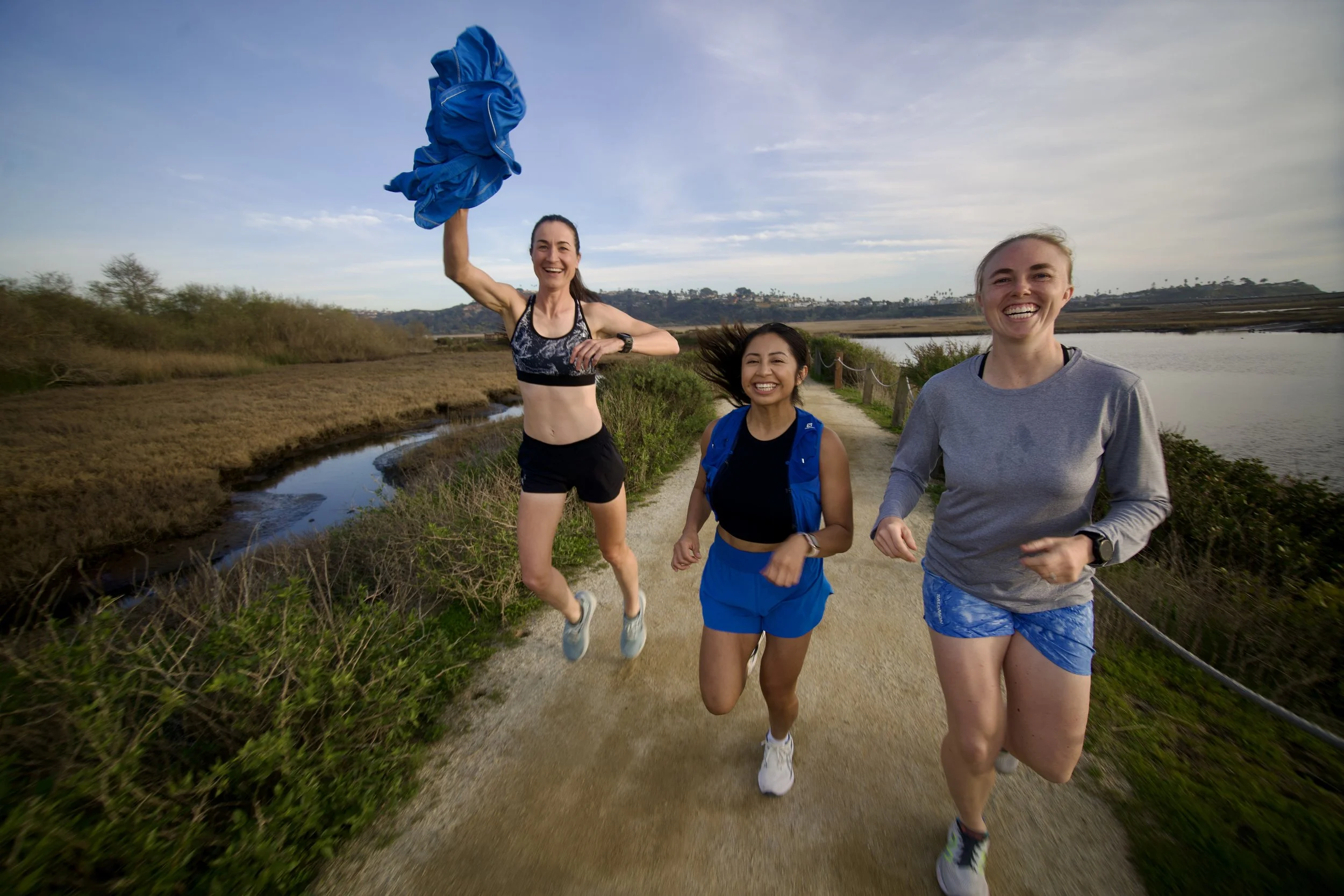 Three women jogging outdoors on a trail near water, with a woman in the middle jumping and holding a blue jacket in the air, all smiling and enjoying a scenic view.