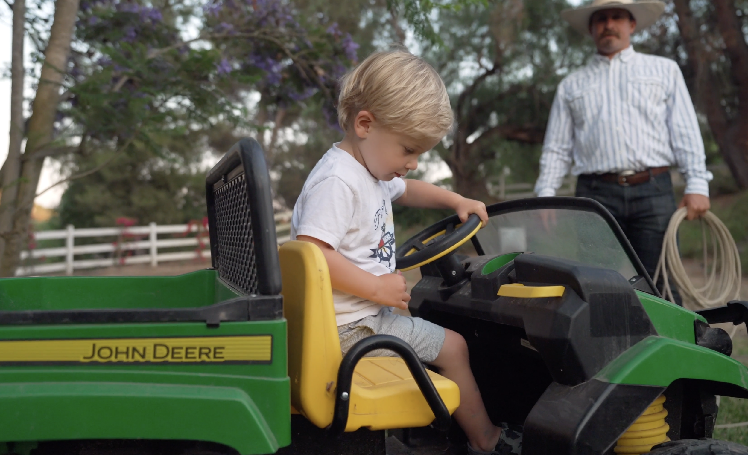 A young boy with blonde hair is sitting in a green John Deere toy tractor, looking down at the steering wheel. An adult with a cowboy hat and striped shirt stands behind him, holding a hose. There are trees and a white fence in the background, suggesting an outdoor setting with a rural or farm-like atmosphere.
