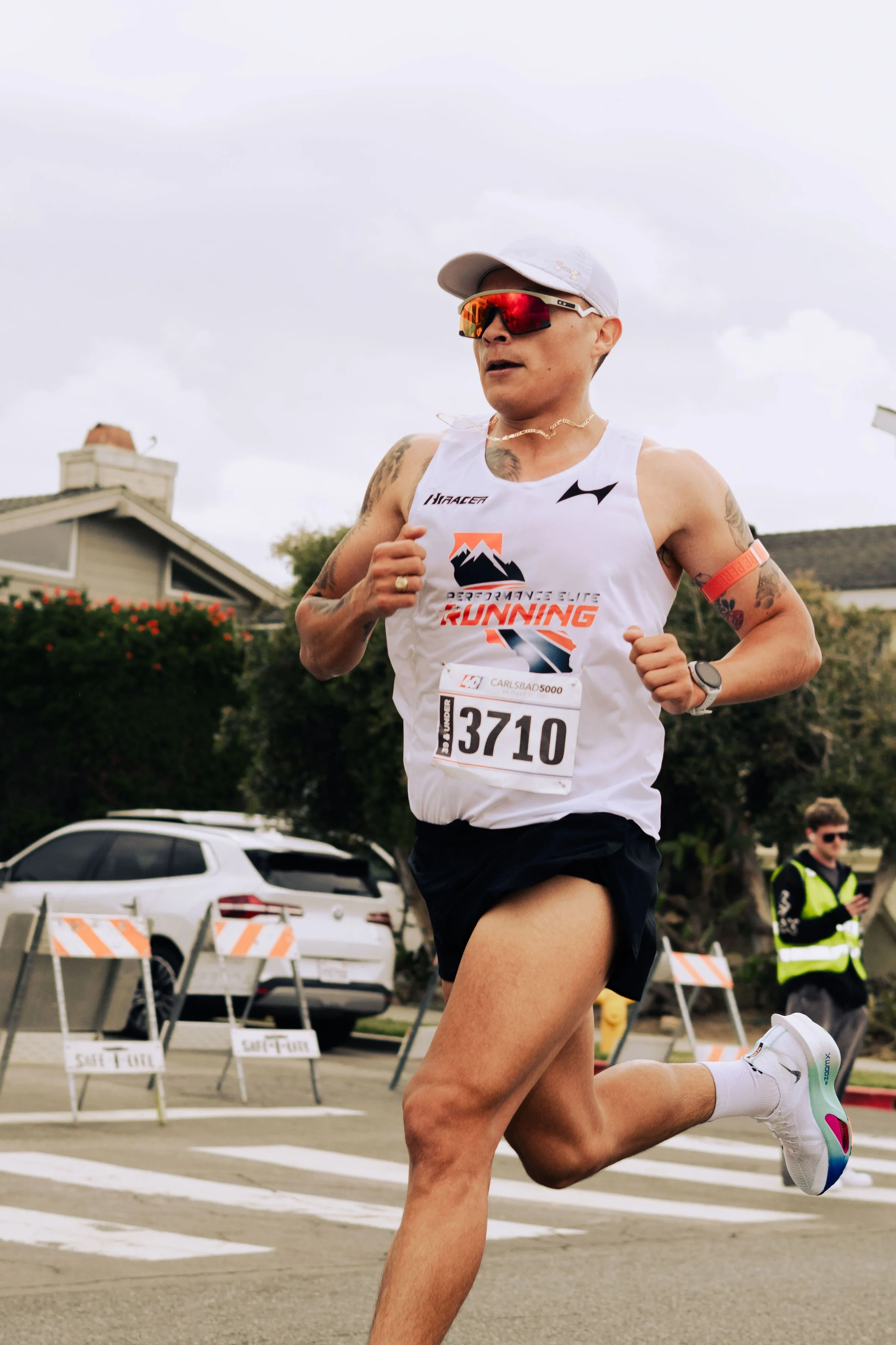 A person running in a race, wearing a white tank top with a logo and the words 'Performance Elite Running,' a bib number 3710, sunglasses, a white cap, and black shorts, with an overcast sky and parking lot in the background.