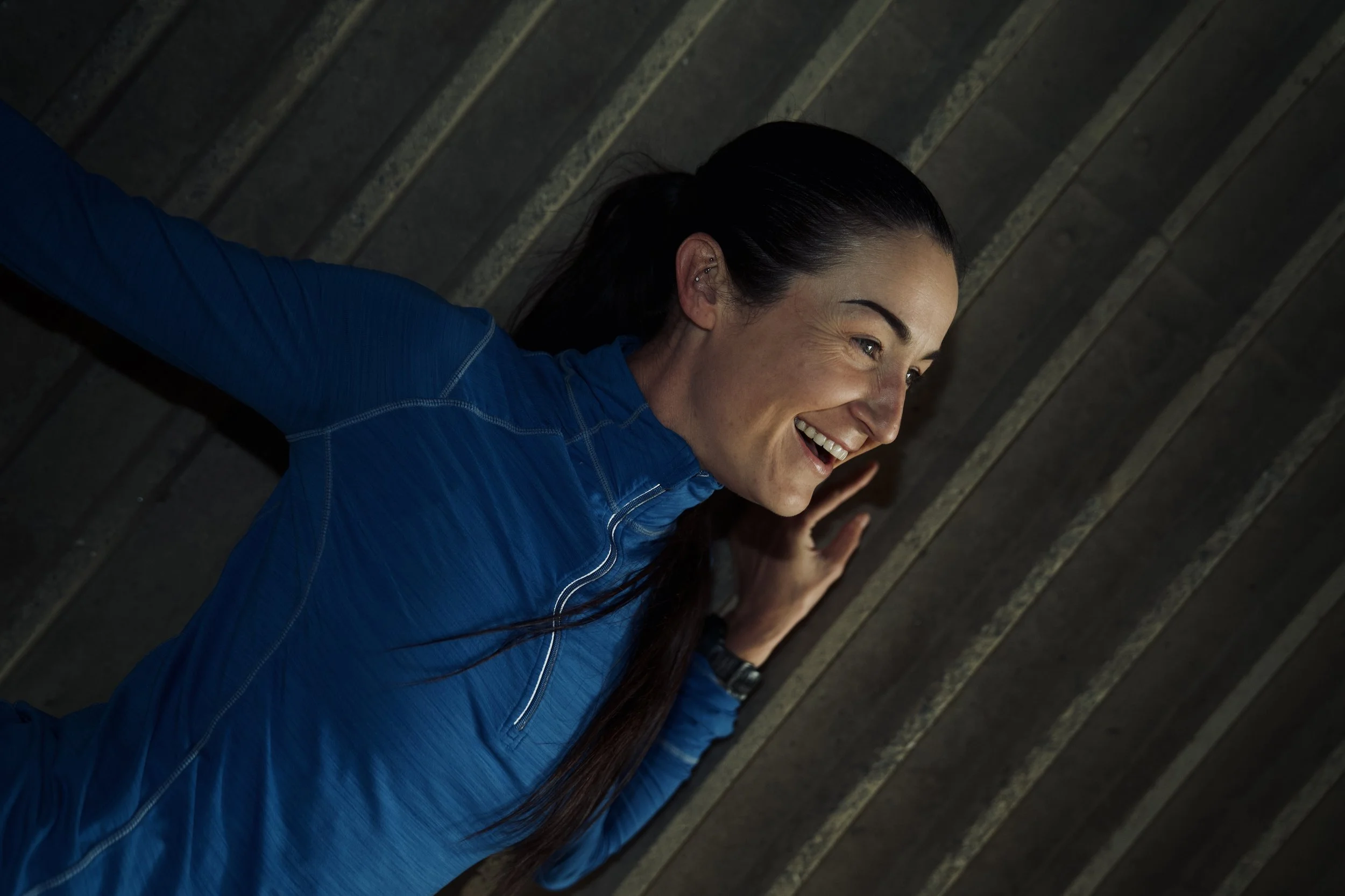 Woman with long dark hair, smiling, wearing a blue jacket, lying on her side on a slanted metal roof.