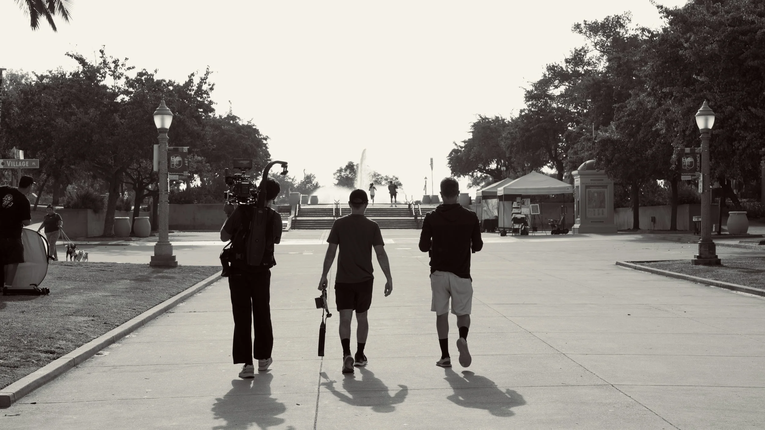 Three young men walking together in an outdoor park setting during daytime, with two holding filming equipment, and a person walking a dog in the background. The scene includes trees, street lamps, tents, and steps leading up to a fountain.