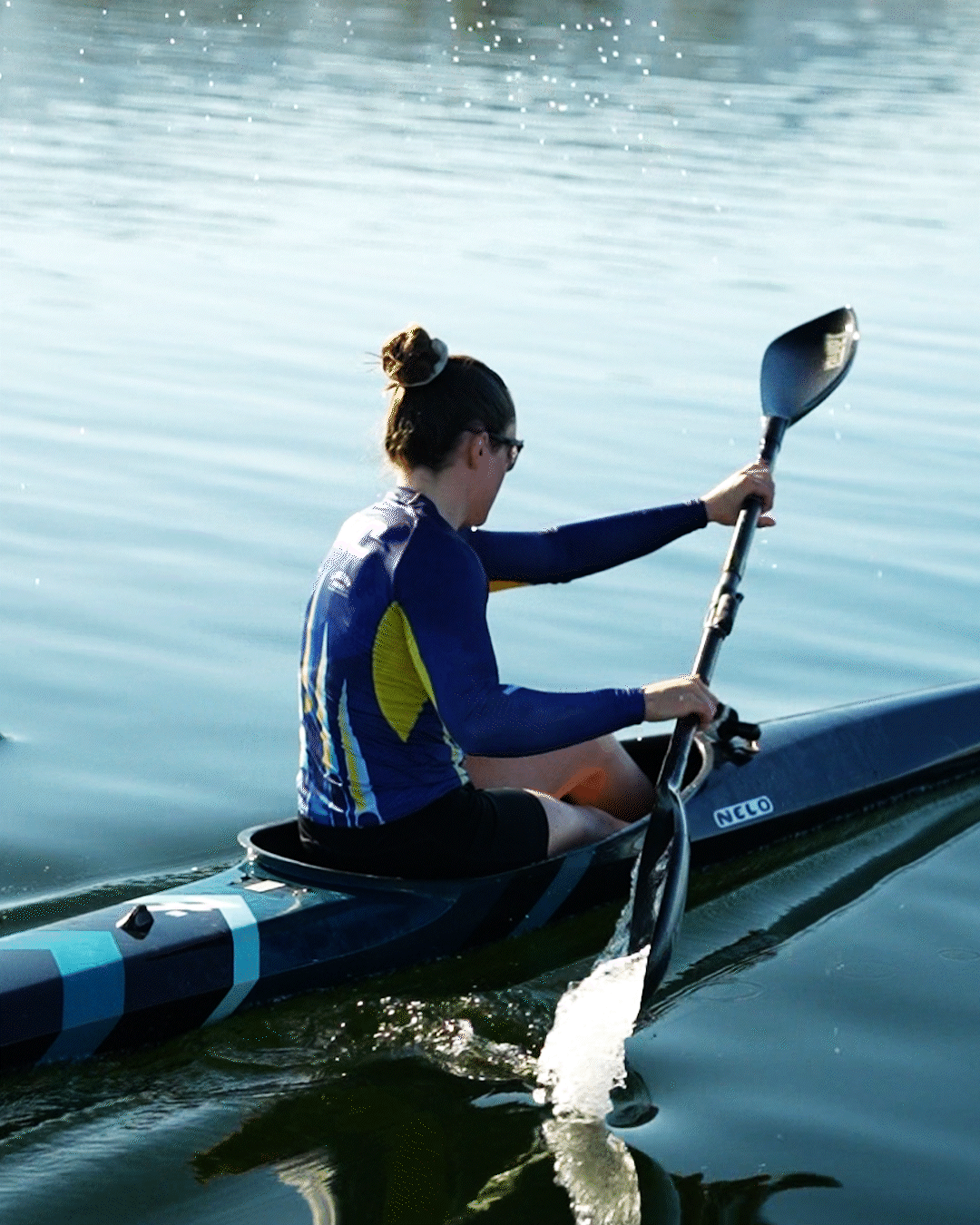A woman kayaking on calm water, wearing a blue long-sleeve shirt, black shorts, and sunglasses, with her hair in a bun, holding a paddle.