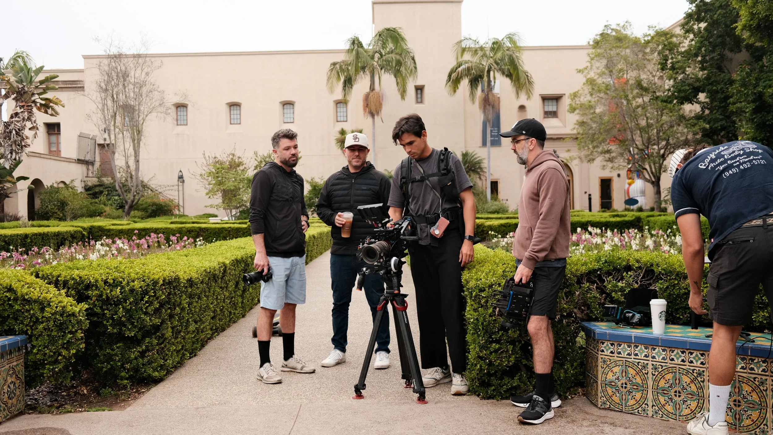 Group of five people working with a camera and filming equipment outdoors in a courtyard with greenery and palm trees.