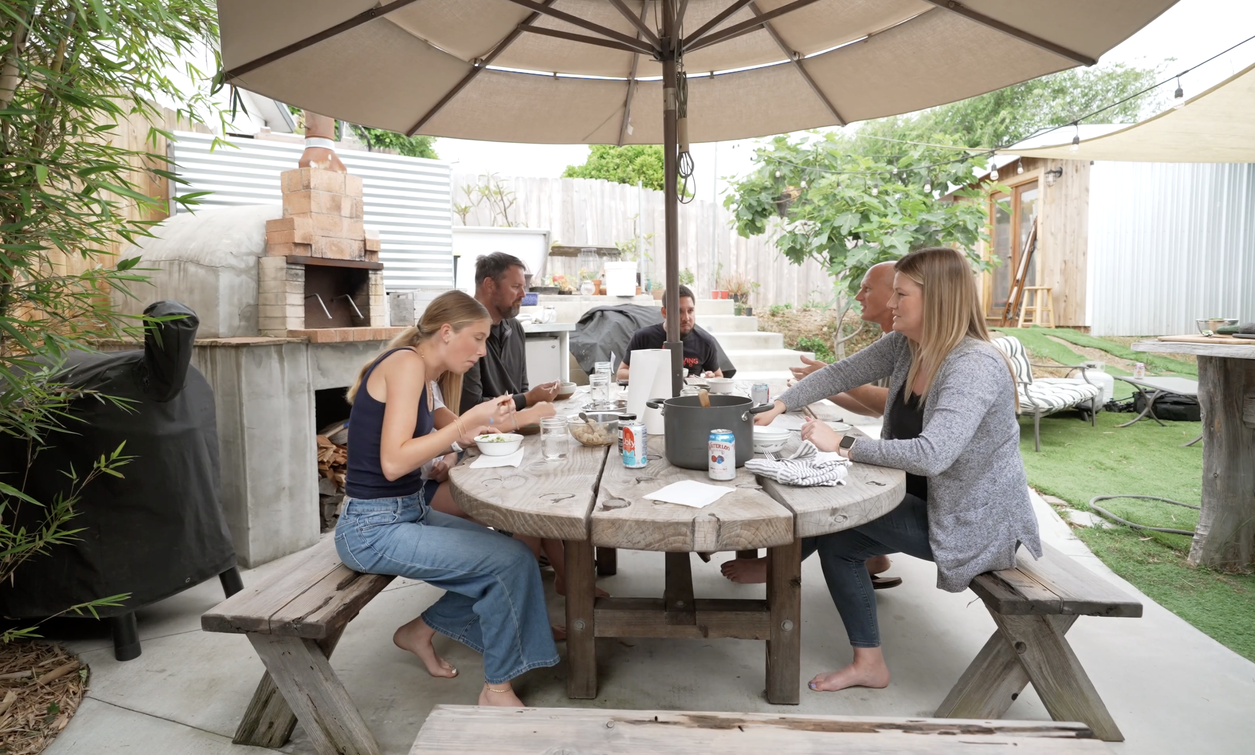 A group of six people sitting at a rustic wooden outdoor dining table under a large beige umbrella in a backyard garden with a brick oven and greenery, having a meal together.