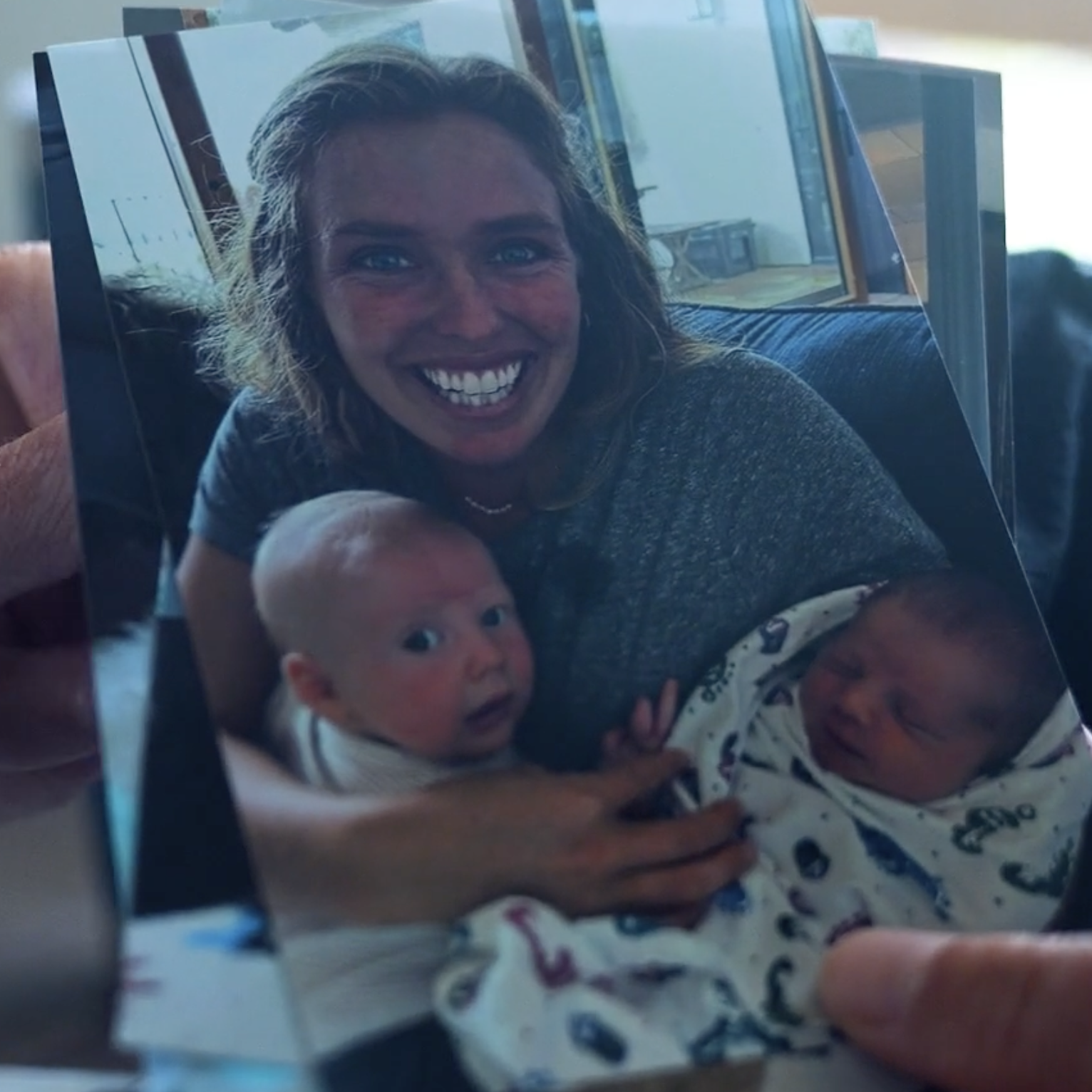 A woman smiling happily while holding two babies, one older and one younger, in front of a window with a bright background.