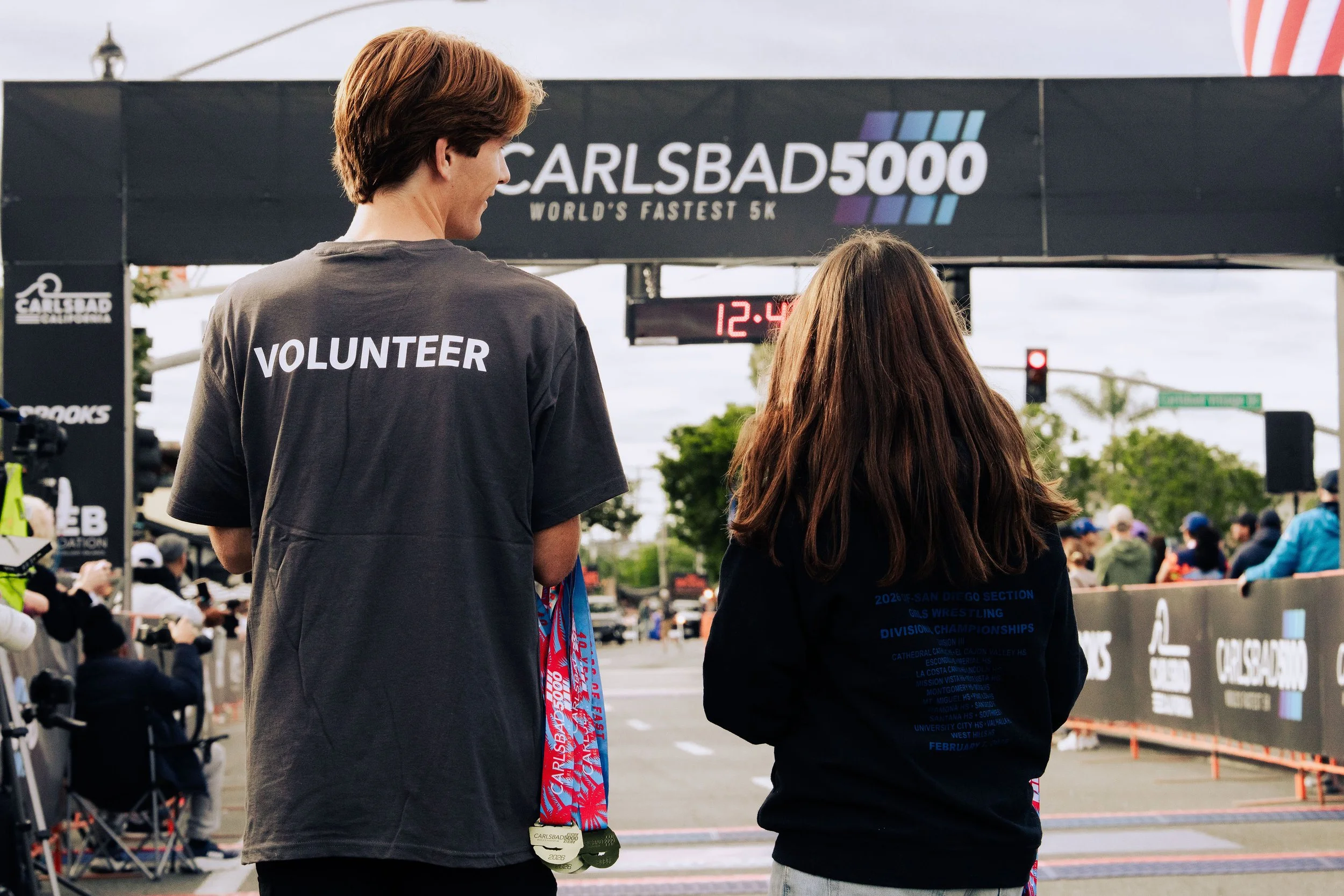 Two people, a man and a woman, standing at a race event with a banner overhead that reads 'CARLSBAD 5000 WORLD'S FASTEST 5K'. The man is wearing a gray 'VOLUNTEER' shirt and has medals hanging around his neck. The woman is wearing a dark hoodie with 