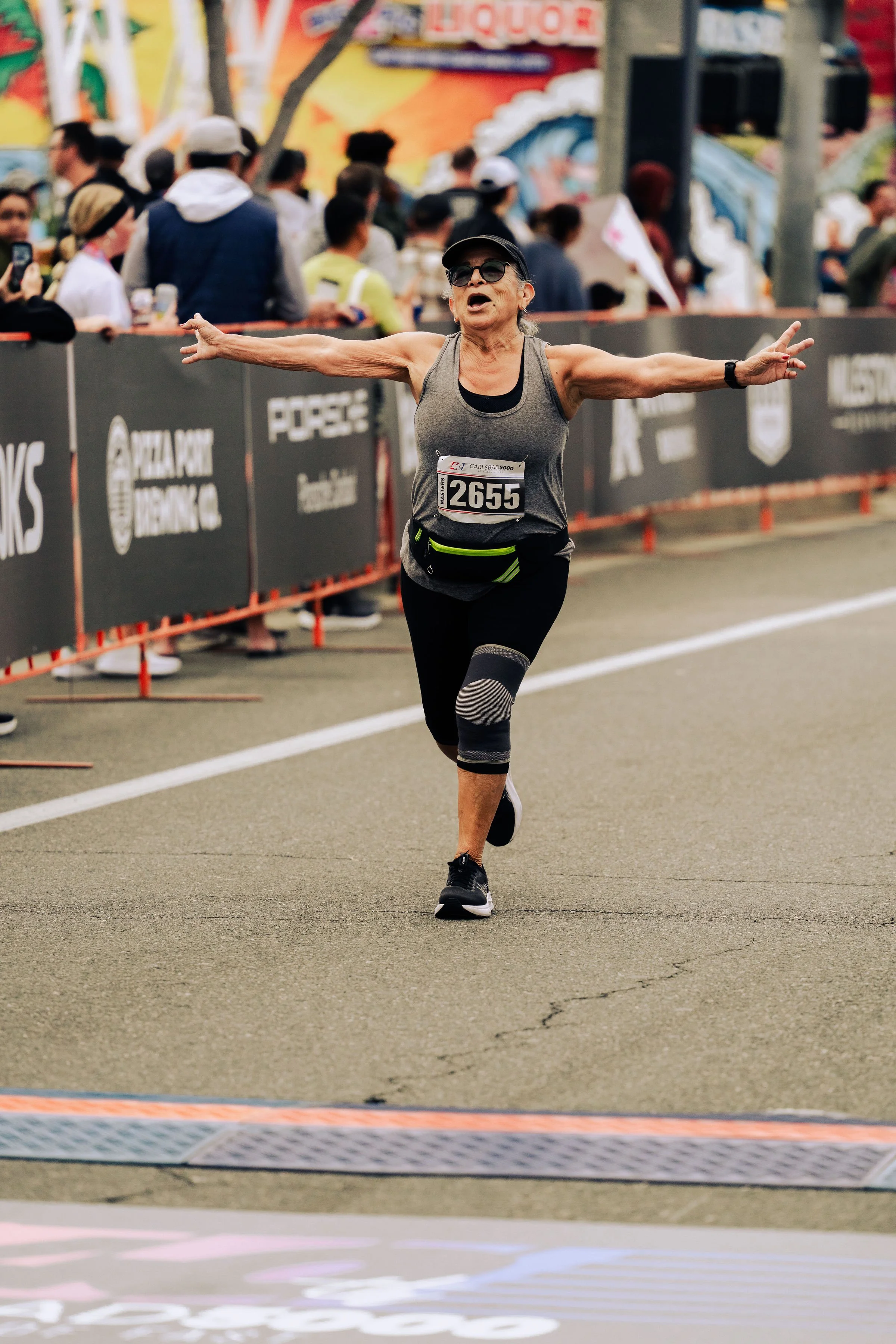 An older woman running in a marathon, wearing a black cap, dark sunglasses, a gray athletic tank top, black leggings with gray knee patches, and a black waist pouch. She has her arms outstretched, appearing happy, with a crowd of spectators and color