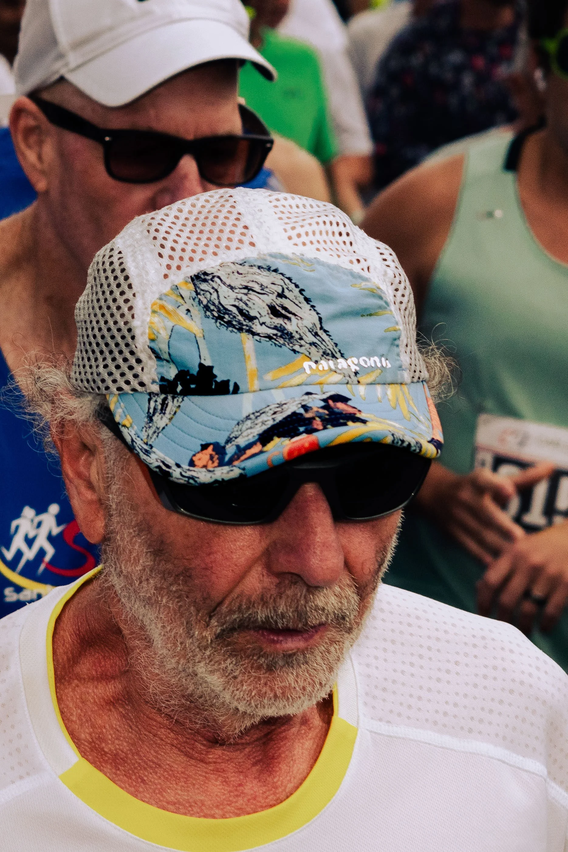 Close-up of an older man with a white beard wearing sunglasses, a white cap, and a white sleeveless athletic shirt with yellow trim, participating in a sporting event.
