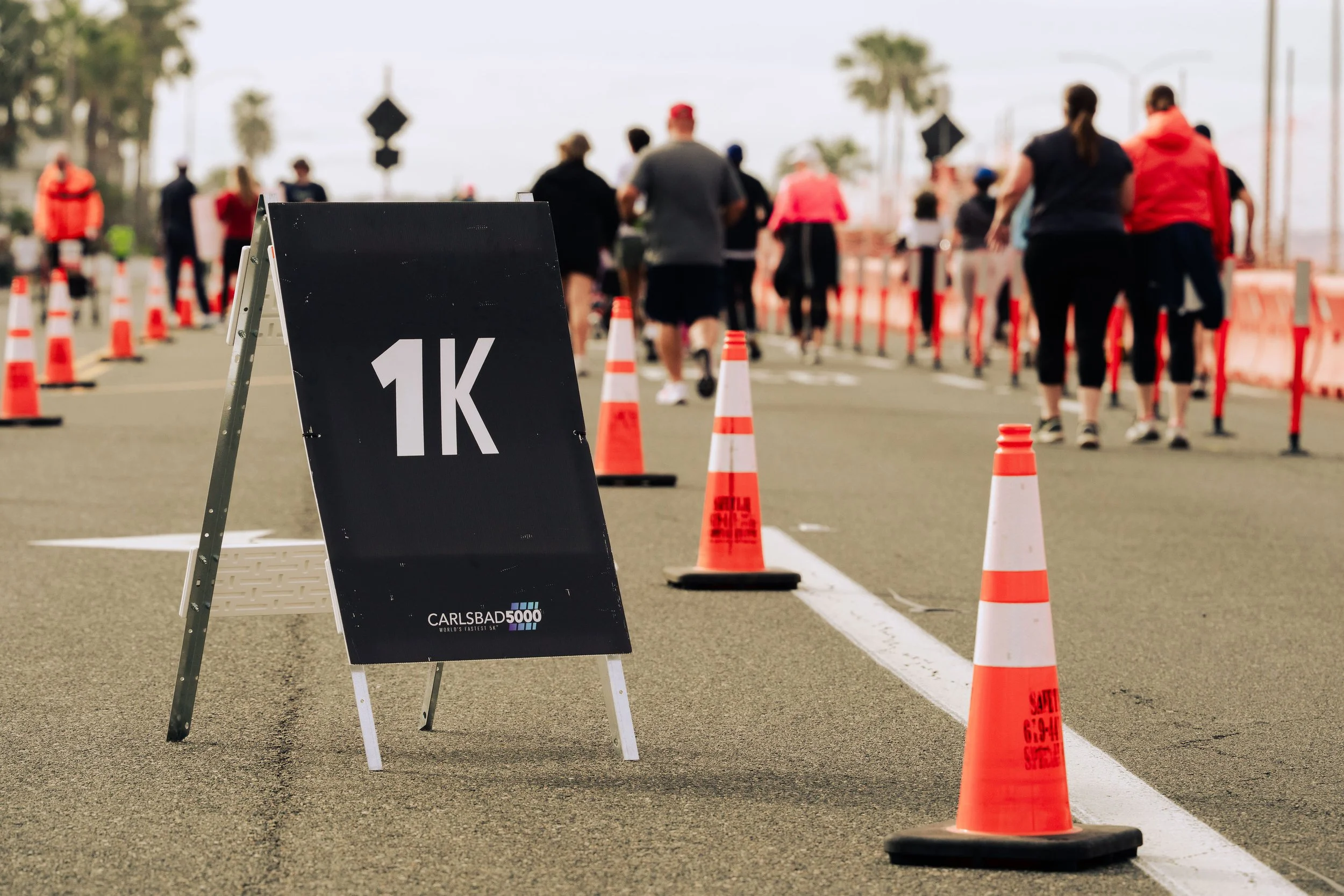 People participating in a 1K run or walk event, lined up behind orange cones on a city street, with a sign in the foreground indicating the 1K distance.