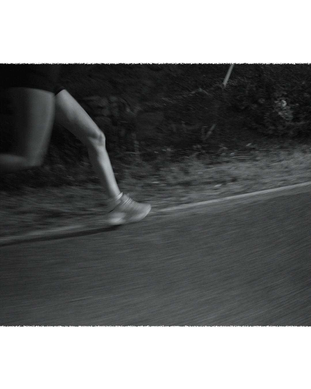Close-up of a person running or walking on a curved asphalt road, with a dirt shoulder and dark surroundings, black and white photo.