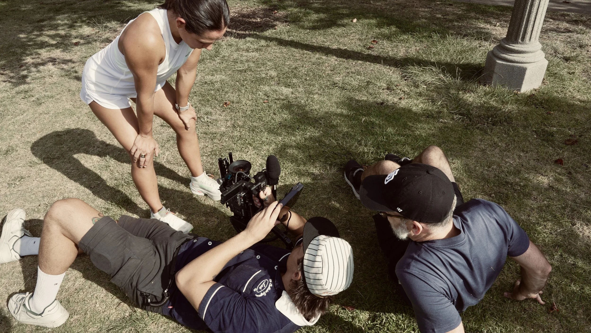 A group of people lying on the grass, with one person standing and others sitting or lying around a professional camera on a stabilizer, on a sunny day in a park-like setting.