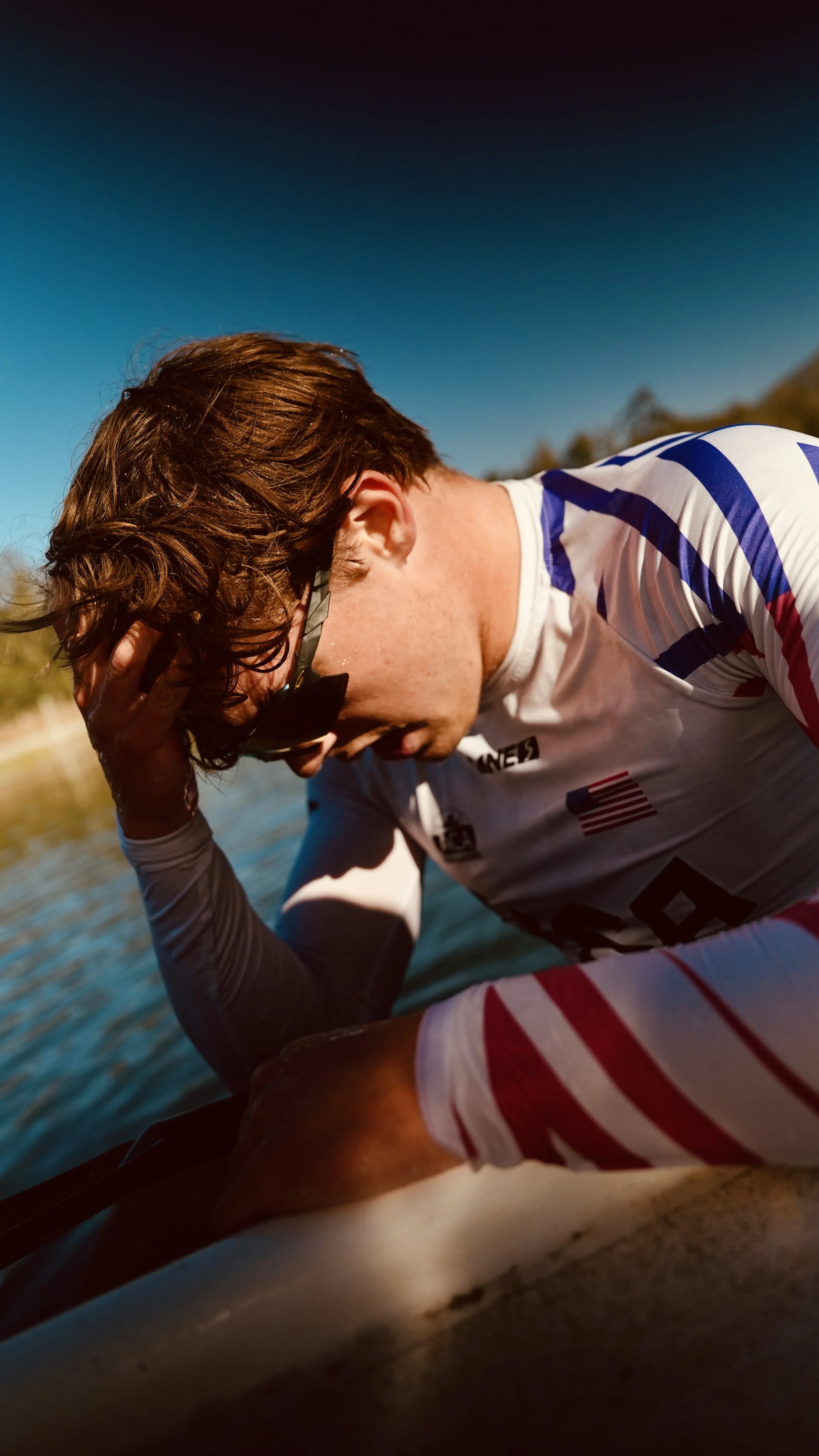 A man wearing sunglasses and a sports jersey is outdoors, leaning on a boat, with water and trees in the background, during daytime.