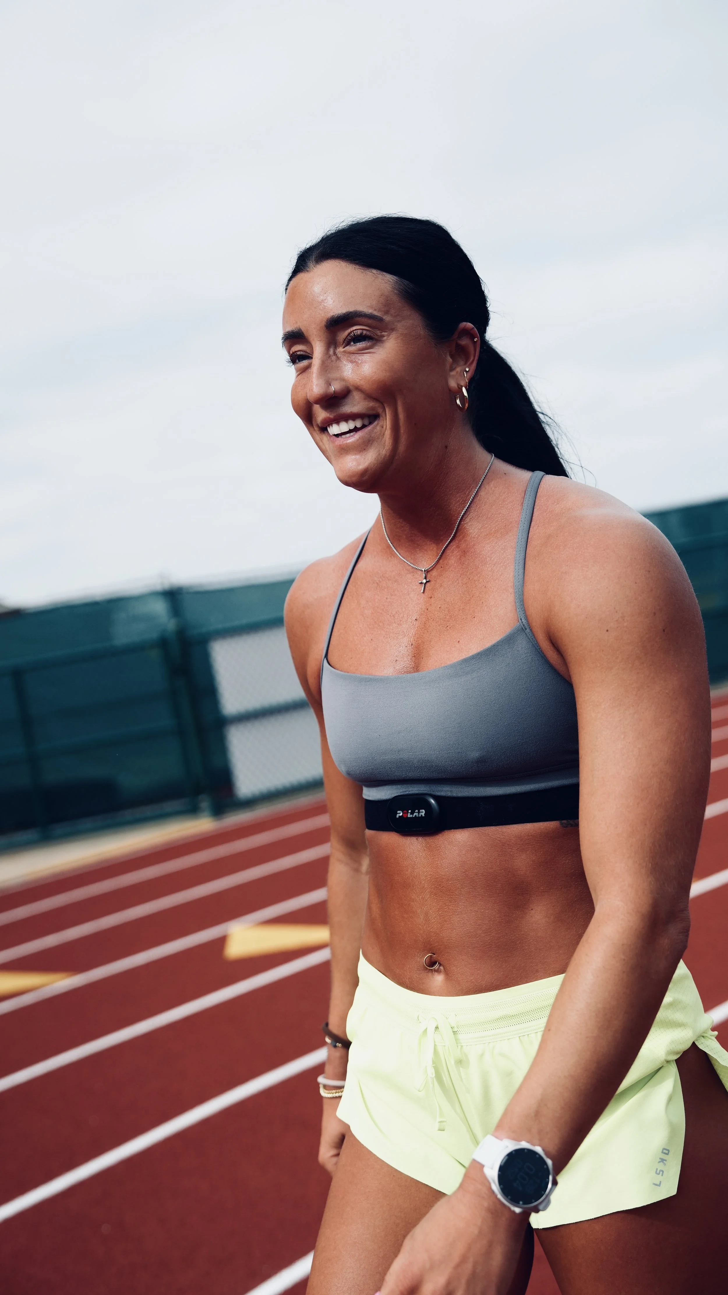 A woman smiling and wearing athletic gear on a running track outdoors, with a fitness device on her waist and a smartwatch on her wrist.