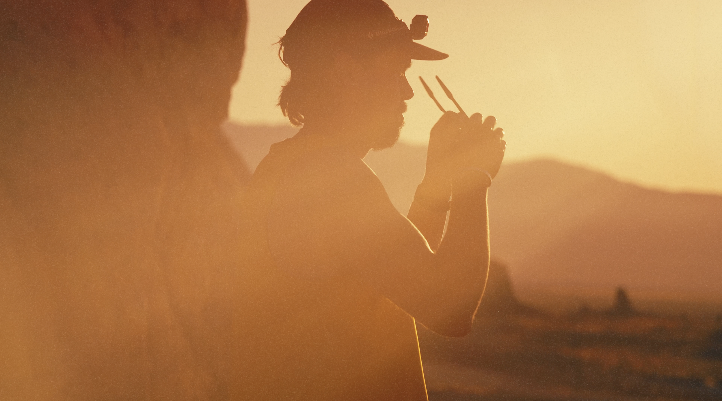 A silhouette of a person wearing a cap and headlamp, with a beard, holding a walkie-talkie or communication device, against a setting sun, possibly outdoors.