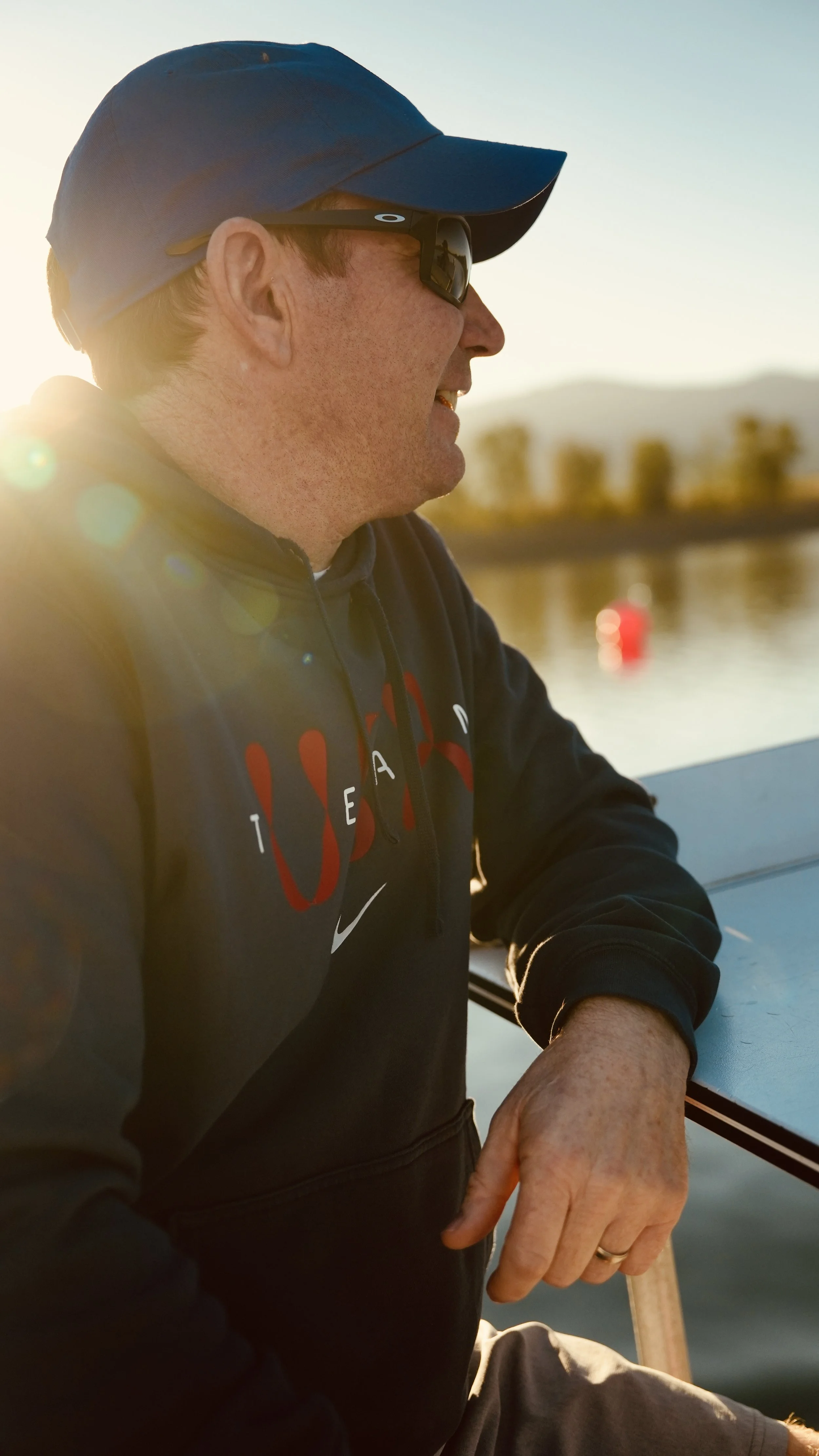 A man wearing sunglasses and a blue cap smiling and looking out at a body of water during sunset, with trees in the background.