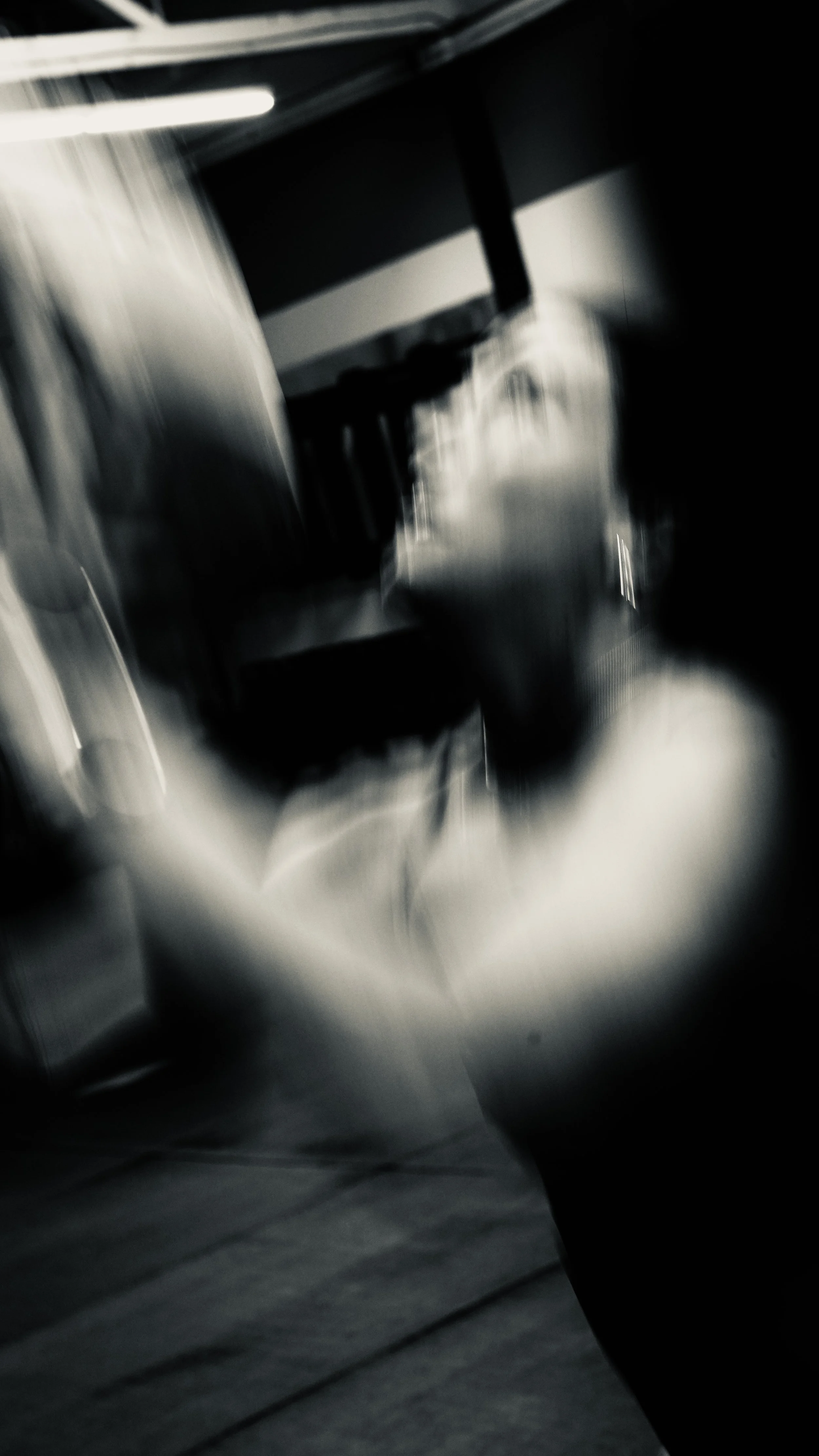 A blurry black-and-white photo of a person standing near a metallic structure, possibly in an industrial or storage setting, with blurred heavy machinery or equipment in the background.