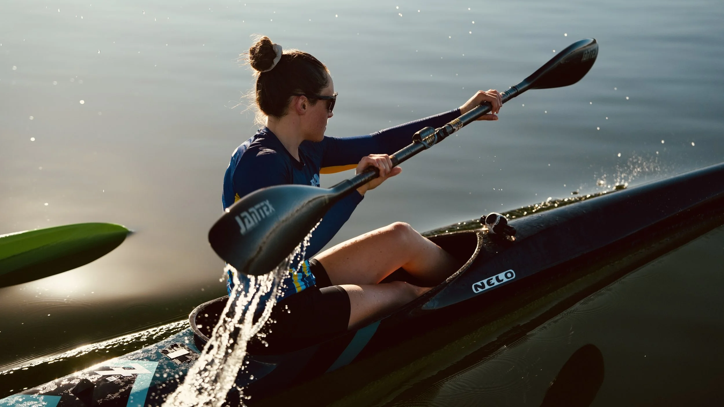 A woman in a kayak paddling on a calm body of water during sunset, wearing sunglasses and a navy blue life vest.