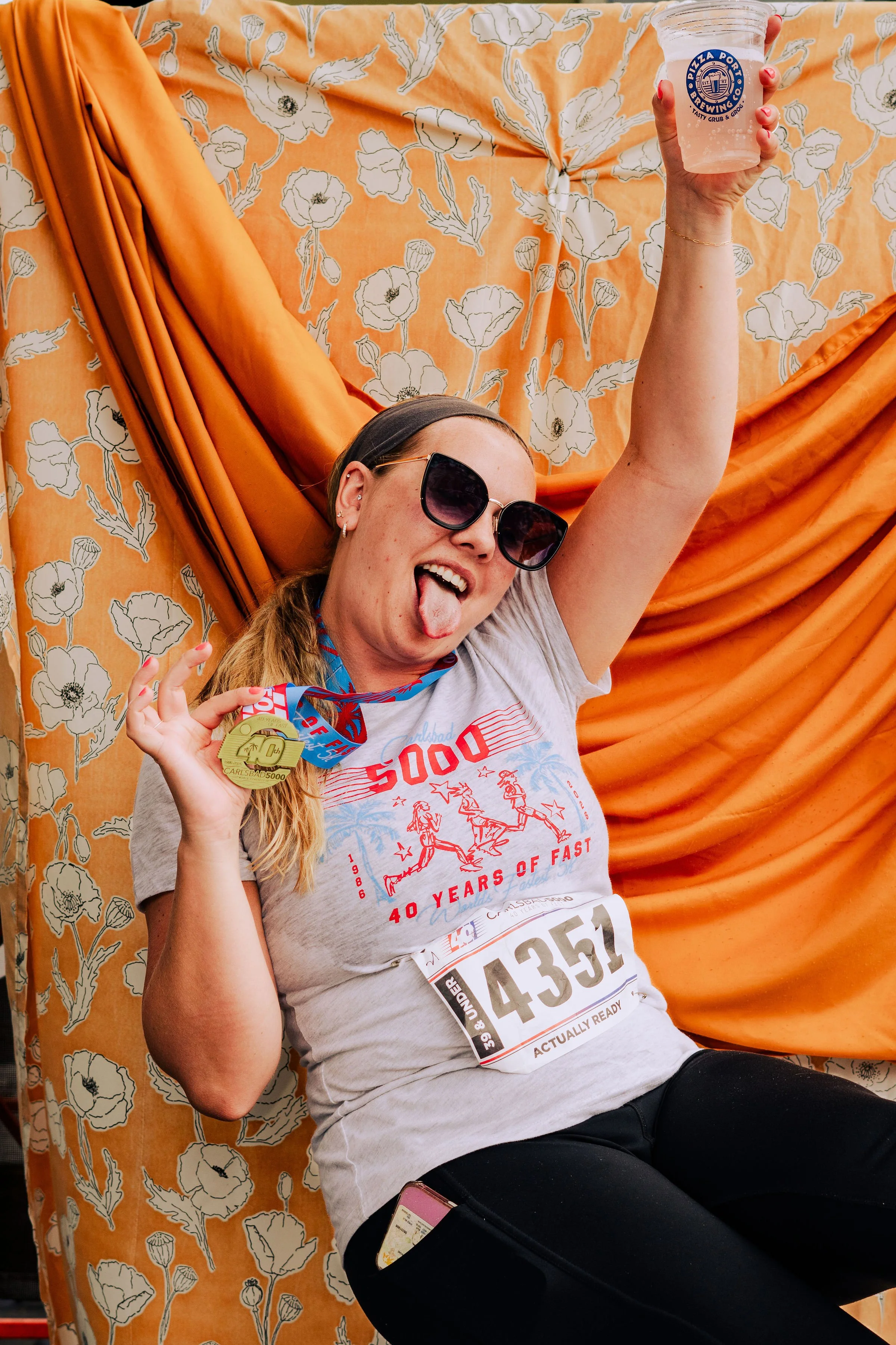 A woman wearing sunglasses and a gray T-shirt with a runner’s bib is holding a plastic cup, celebrating after completing a race. She is smiling and sticking out her tongue, and is wearing medals around her neck. The background is decorated with orang