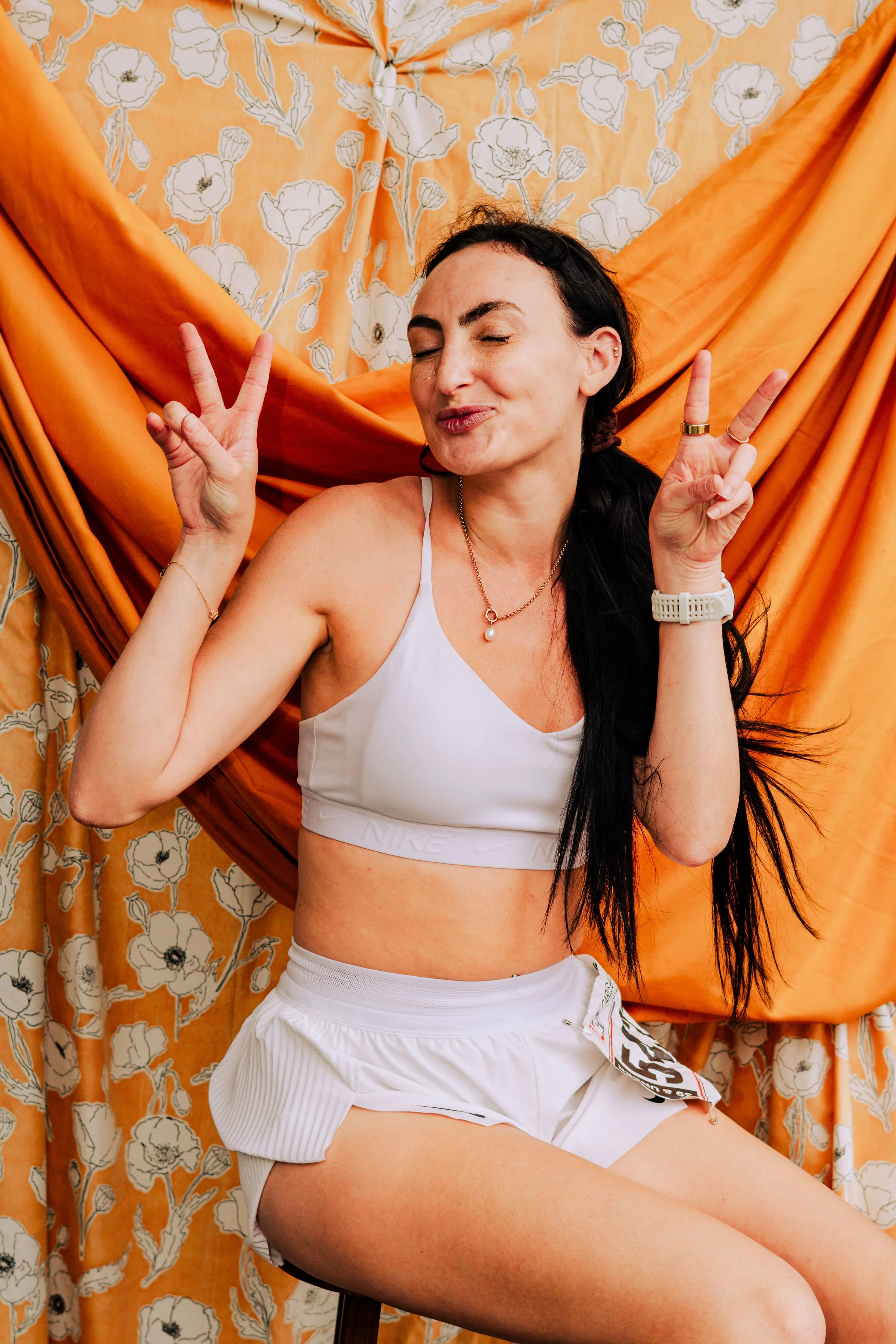 A woman with long dark hair wearing a white sports bra and white shorts sitting in front of orange floral patterned drapes. She is making a peace sign with both hands and has a content expression with her eyes closed.