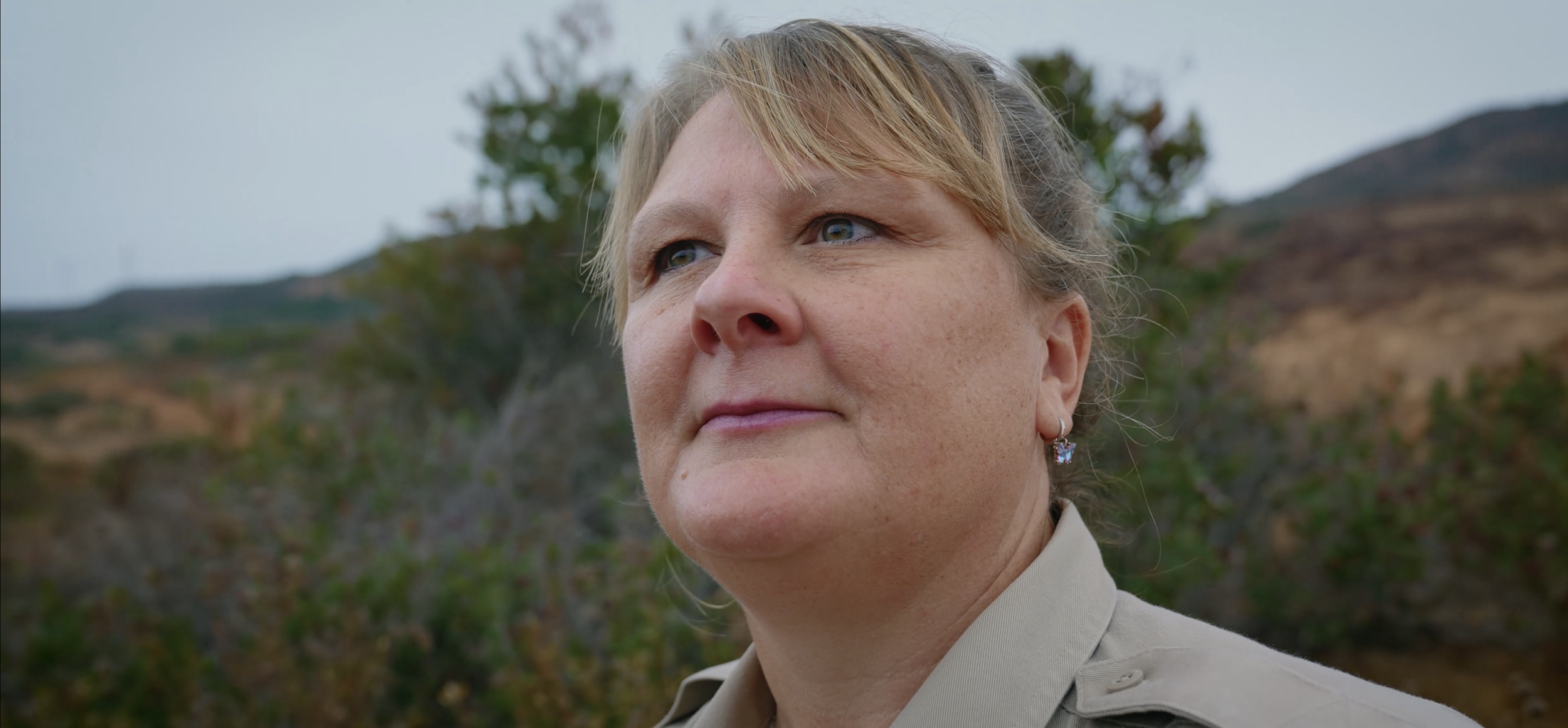Close-up of a woman with short blonde hair and blue eyes, wearing a beige jacket and floral earrings, standing outdoors with mountains and trees in the background.