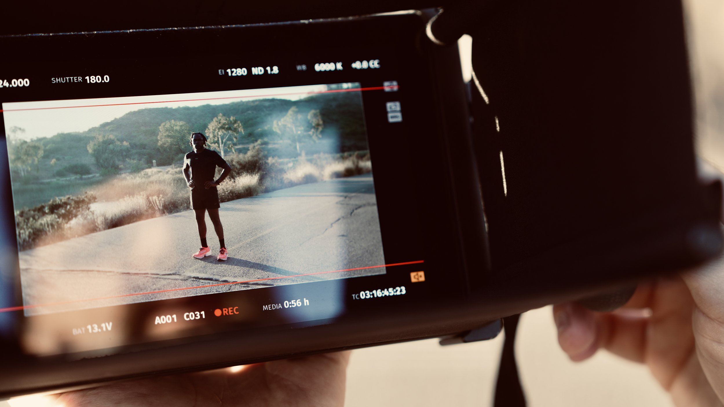 A person holding a camera captures a photo of a man standing on a rural road with trees and hills in the background.