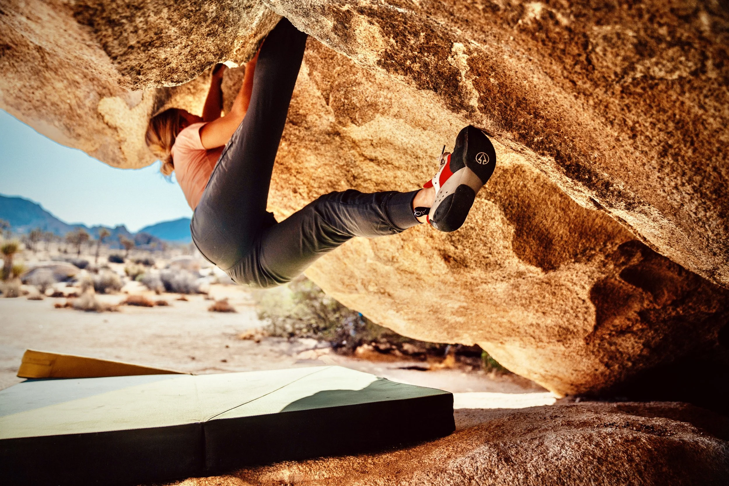 A woman bouldering outdoors on a large rock formation during daytime, wearing climbing shoes and designated workout pants, reaching for a handhold.