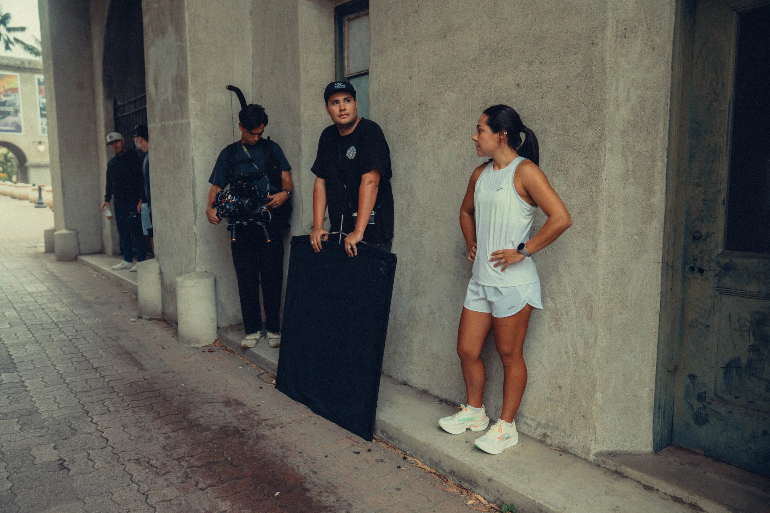 A woman in athletic clothing standing against a wall with her hands on her hips. Two men are standing nearby, one holding a camera and another looking at his phone. There are two other men in the background near an archway.