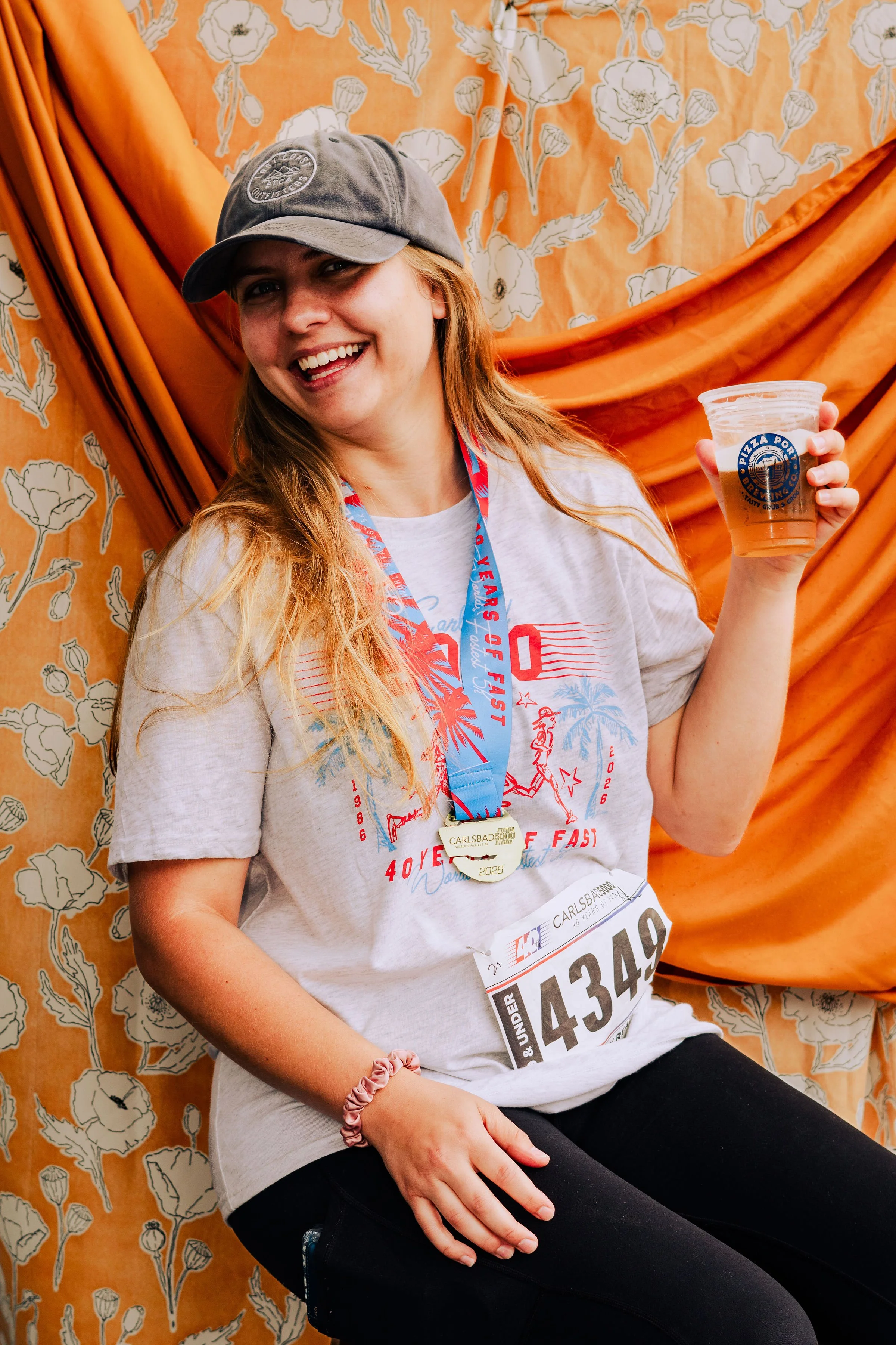 A young woman with long red hair wearing a gray baseball cap, white T-shirt, black leggings, and a pink scrunchie, smiling while holding a plastic cup of beer. She has a running bib with the number A4349 and a medal around her neck. The background fe