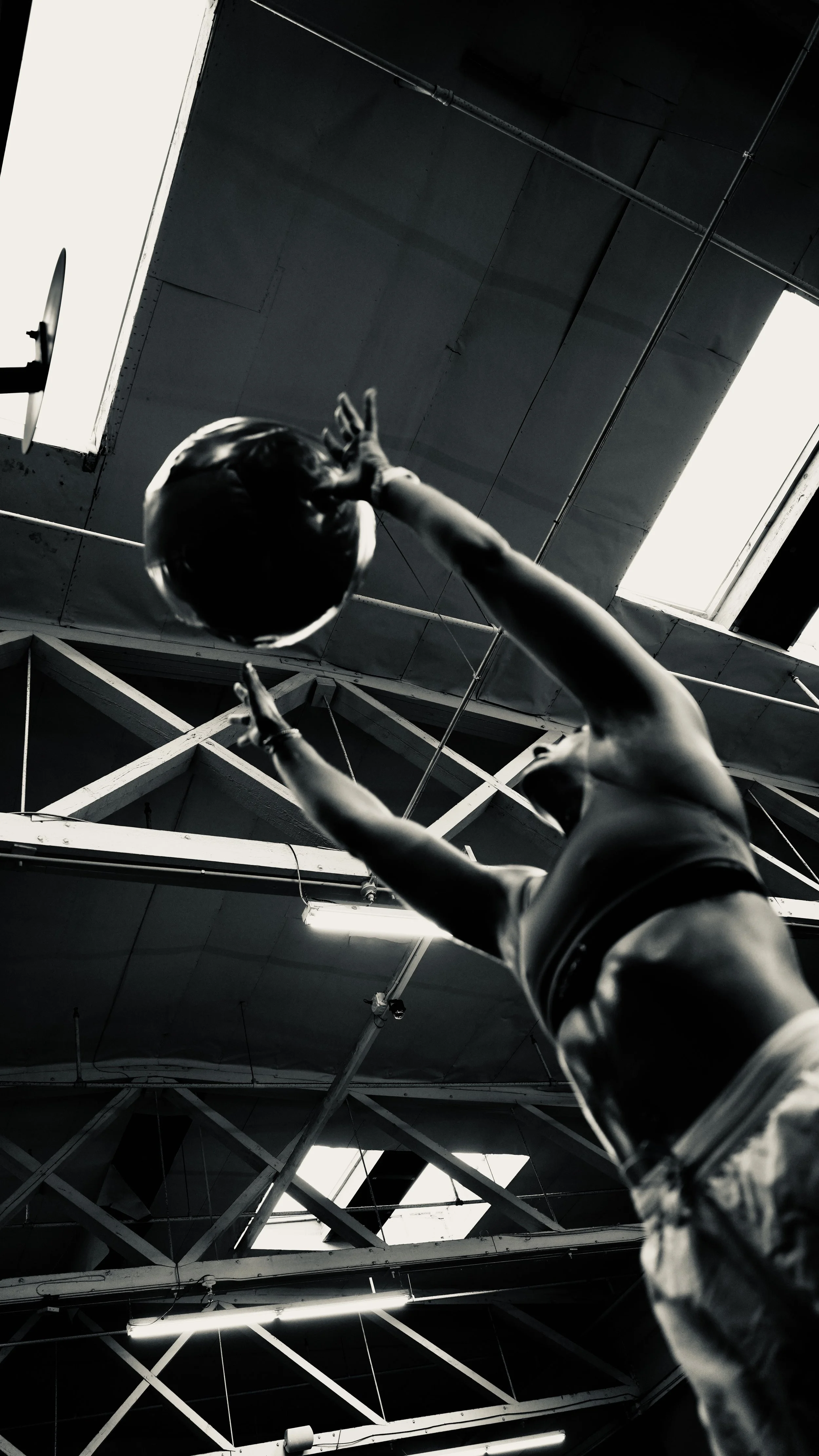 A mannequin dressed in sportswear, with a cap, reaching upward with both hands, in an indoor industrial space with exposed ceiling pipes and a large skylight.