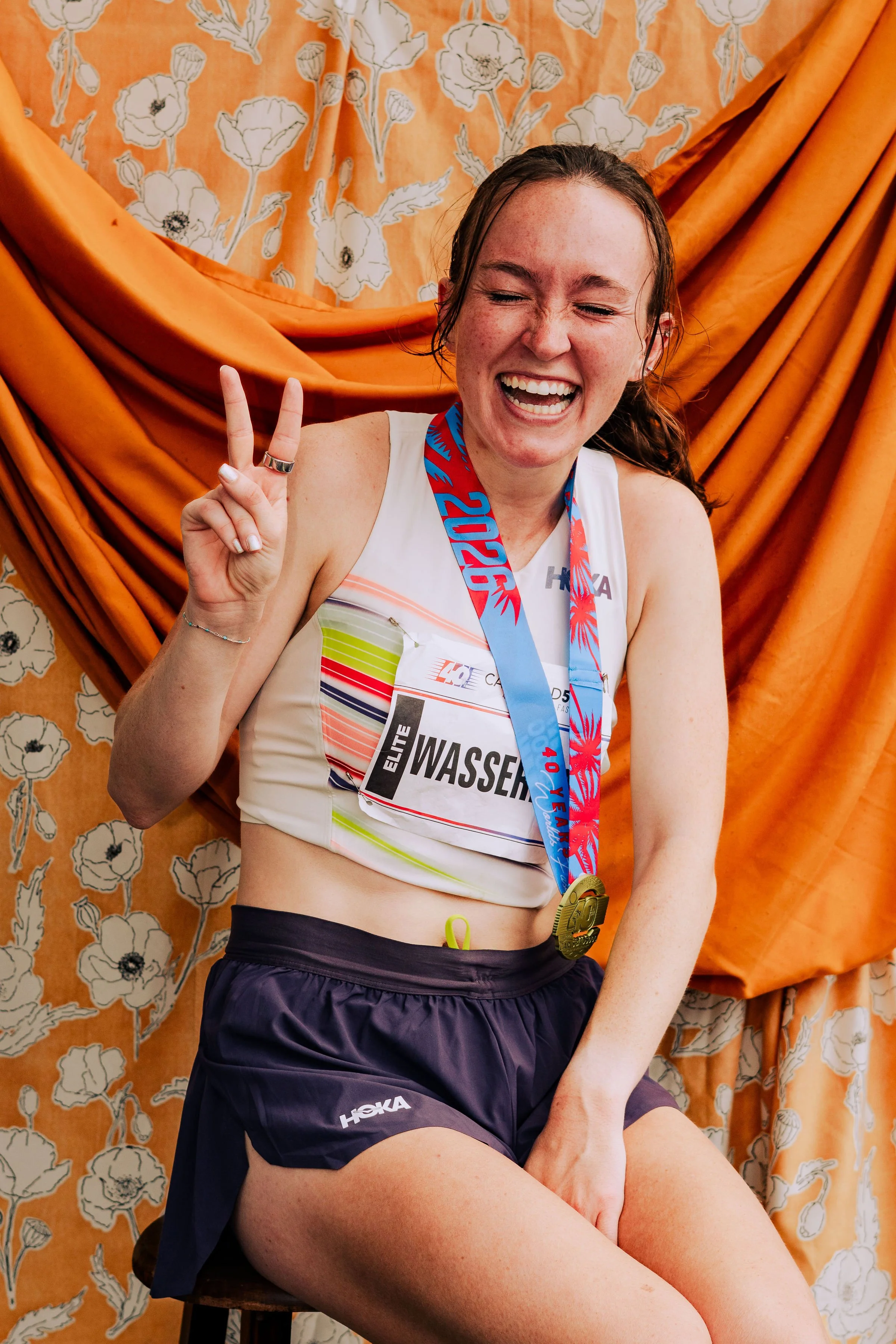 A young woman sits on a stool after a race, smiling and making a peace sign with her fingers. She is wearing a white athletic tank top with colorful stripes, black shorts, and a medal around her neck. The background features orange and floral pattern