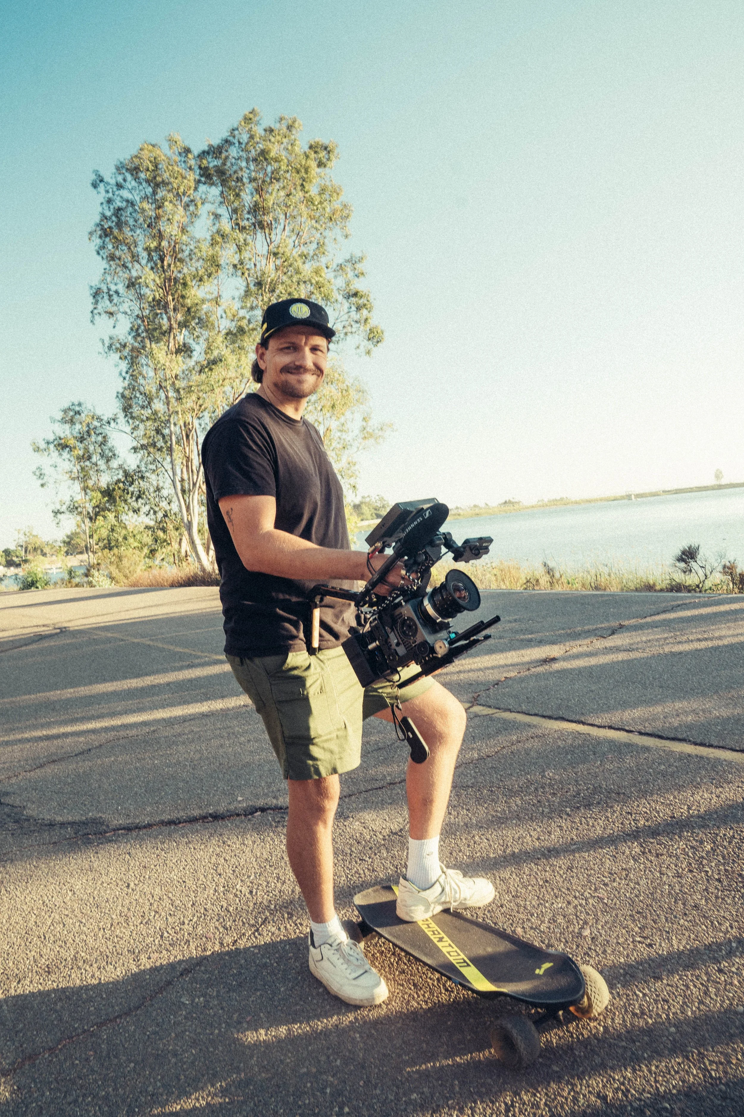 A man smiling while standing outdoors on a skateboard, holding a video camera, with a large tree and water body in the background during daytime.
