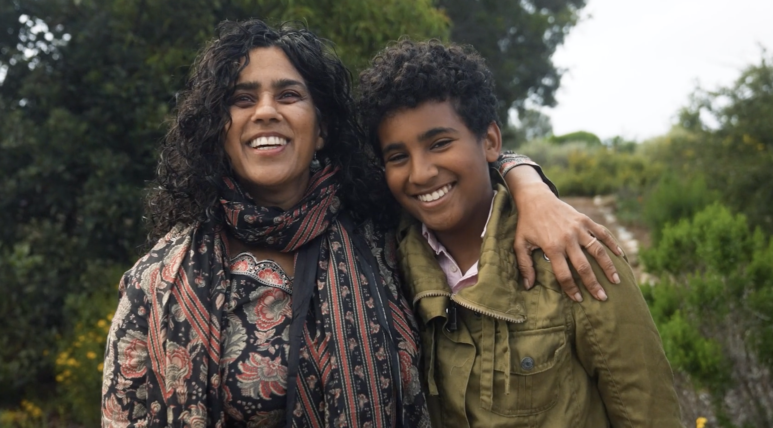 A woman with curly dark hair and a patterned scarf is smiling and hugging a young boy with short curly hair, both outdoors with green trees and foliage in the background.