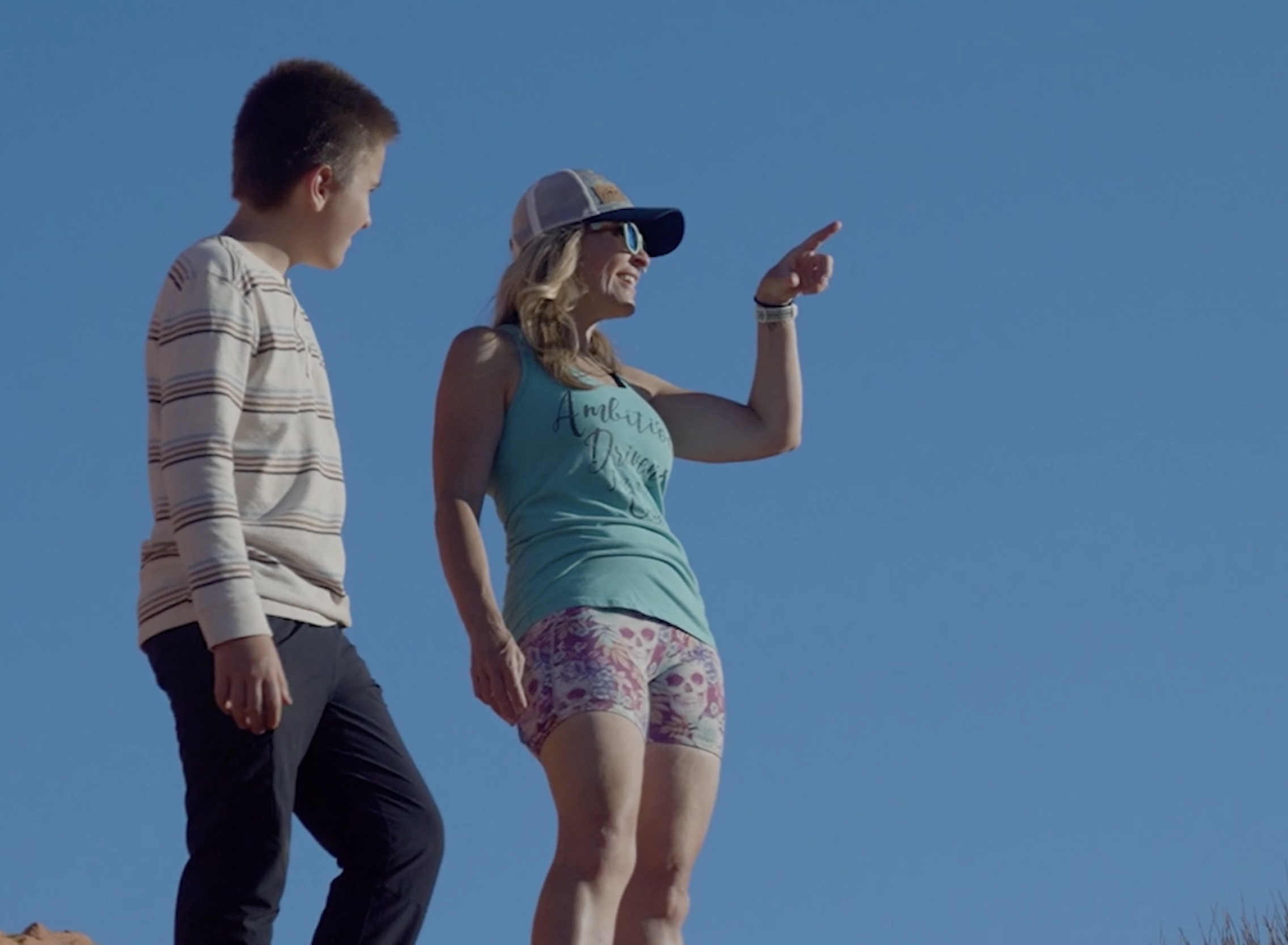 A woman and a young man standing outside under a clear blue sky, holding hands. The woman is wearing a light blue tank top, patterned shorts, sunglasses, and a baseball cap, while the young man is dressed in a striped long-sleeve shirt and dark pants, smiling and looking at each other.