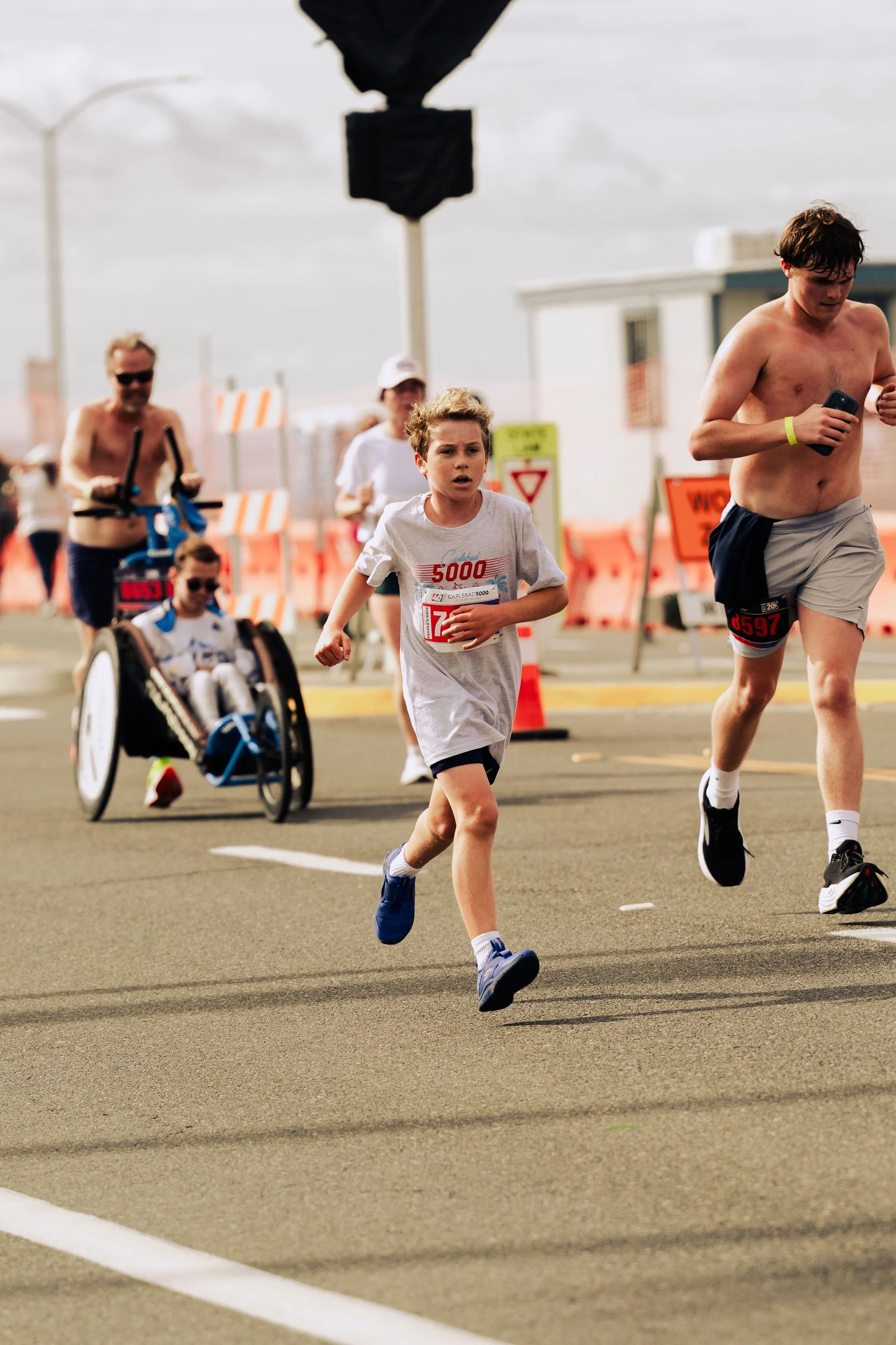 A group of people participating in a running race, with a young boy in the foreground and several adults, including one in a wheelchair, in the background. The scene takes place outdoors on a road with road signs and barriers.
