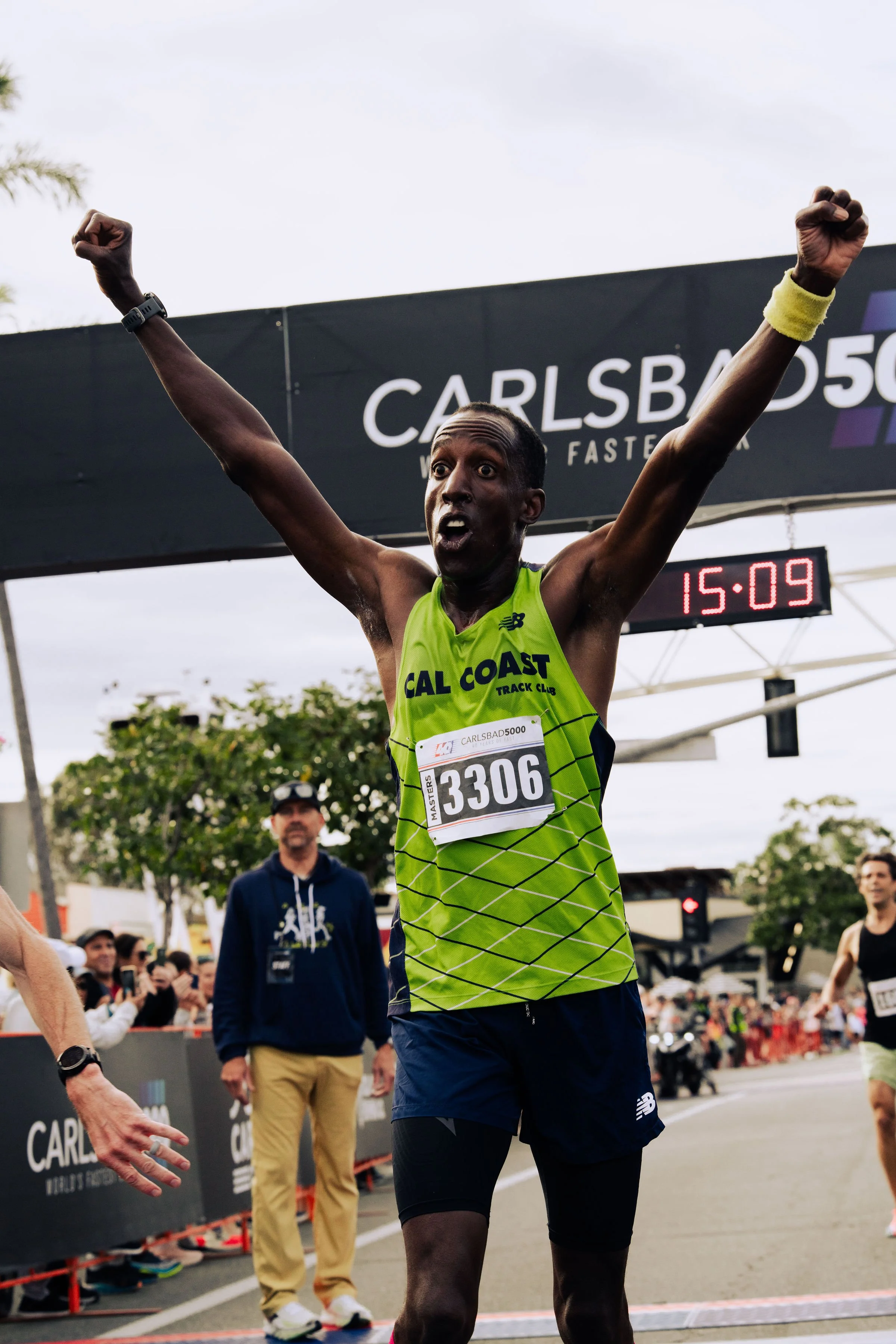 A male marathon runner crossing the finish line with arms raised in victory during a race at Carlsbad, wearing a neon green tank top and black shorts. The time on the digital clock shows 15:09.
